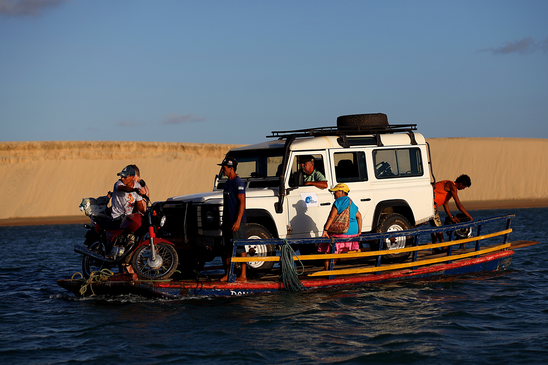A Land Rover, a motorcycle, and some people are seen on a small barge, in the water. On the background a dune. 
