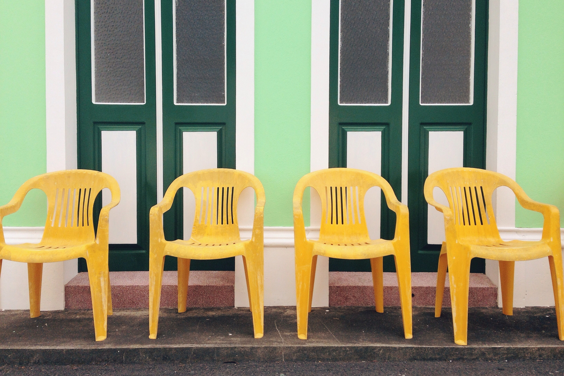 Four yellow plastic chairs neatly arranged against a green wall with green doors. 
