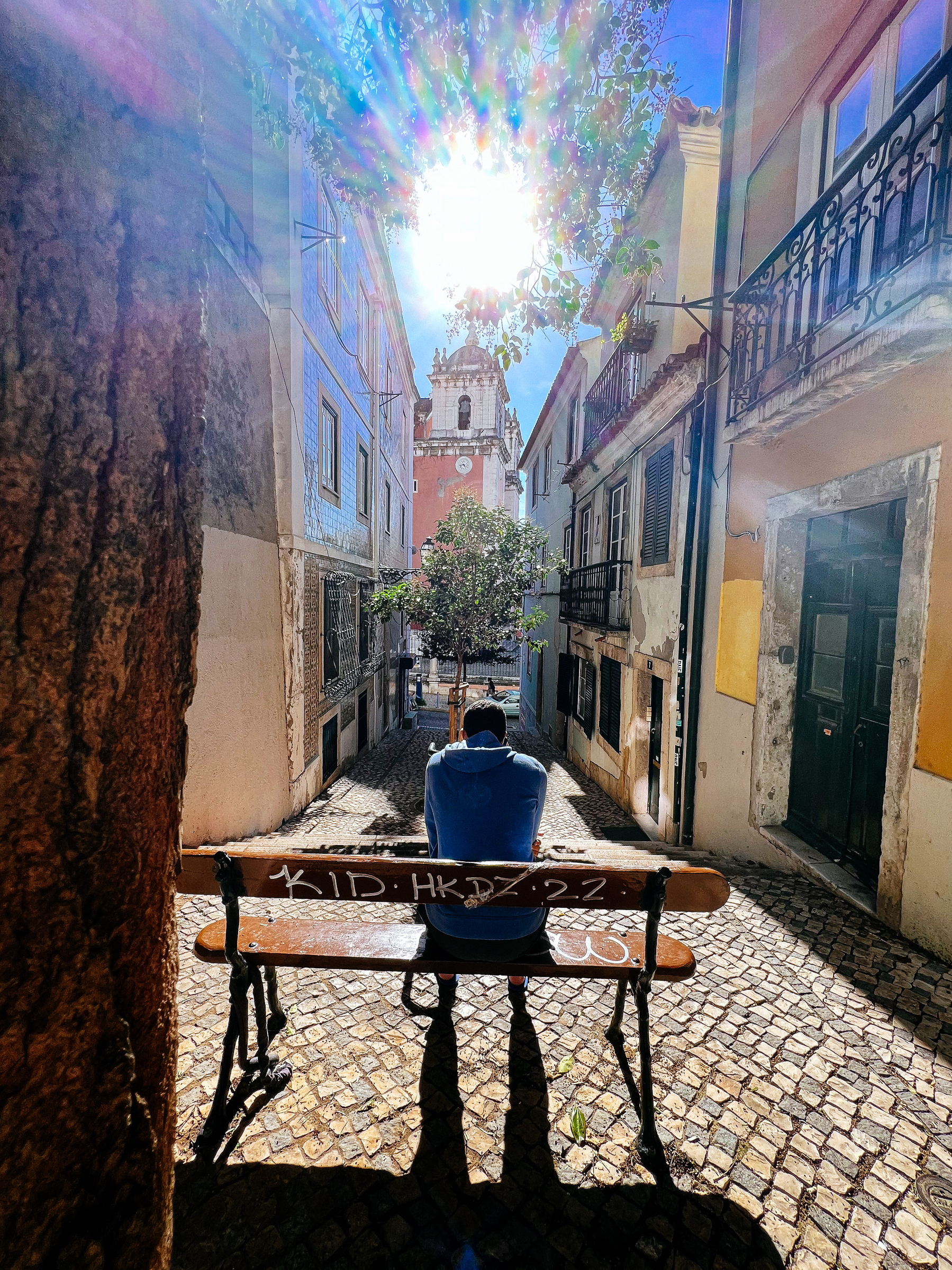 A man sits on a bench, in an alley. On the background is a church, with a burst of light on top 