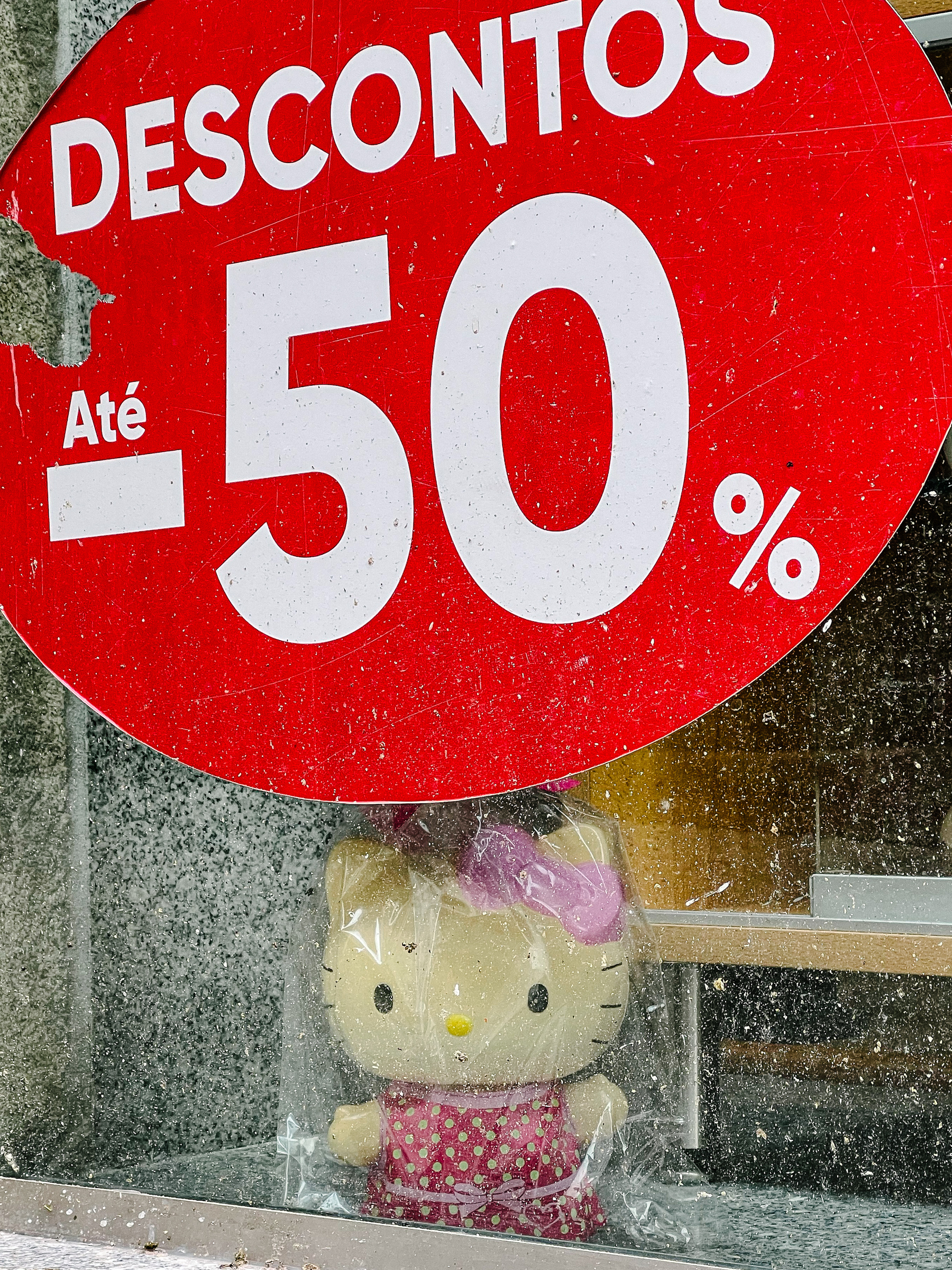 A sad looking Hello Kitty doll sits on a dirty shop window, with a big red sign advertising 50% discounts. 