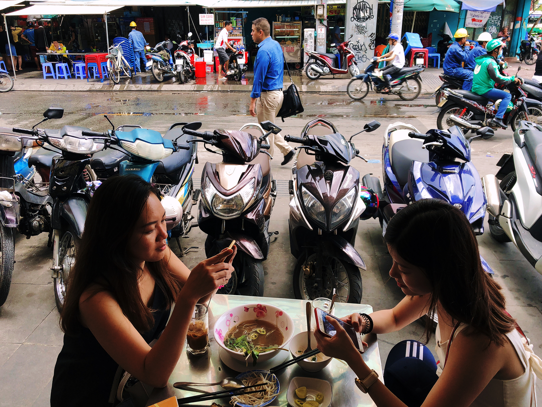 two girls eat a bowl of pho in Vietnam, motorcycles and people in the background