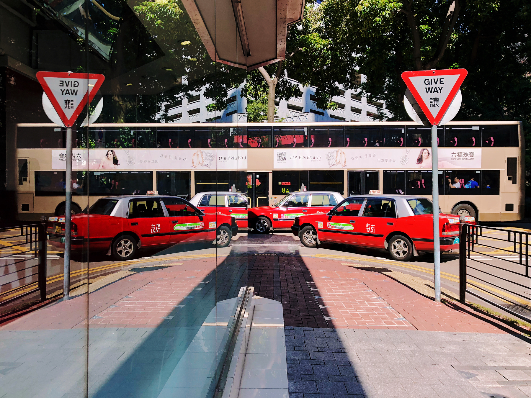 A bus and two taxis are reflected on a window, creating something that’s very close to symmetry. A sign, also reflected, says “Give Way”, in English and Chinese. 
