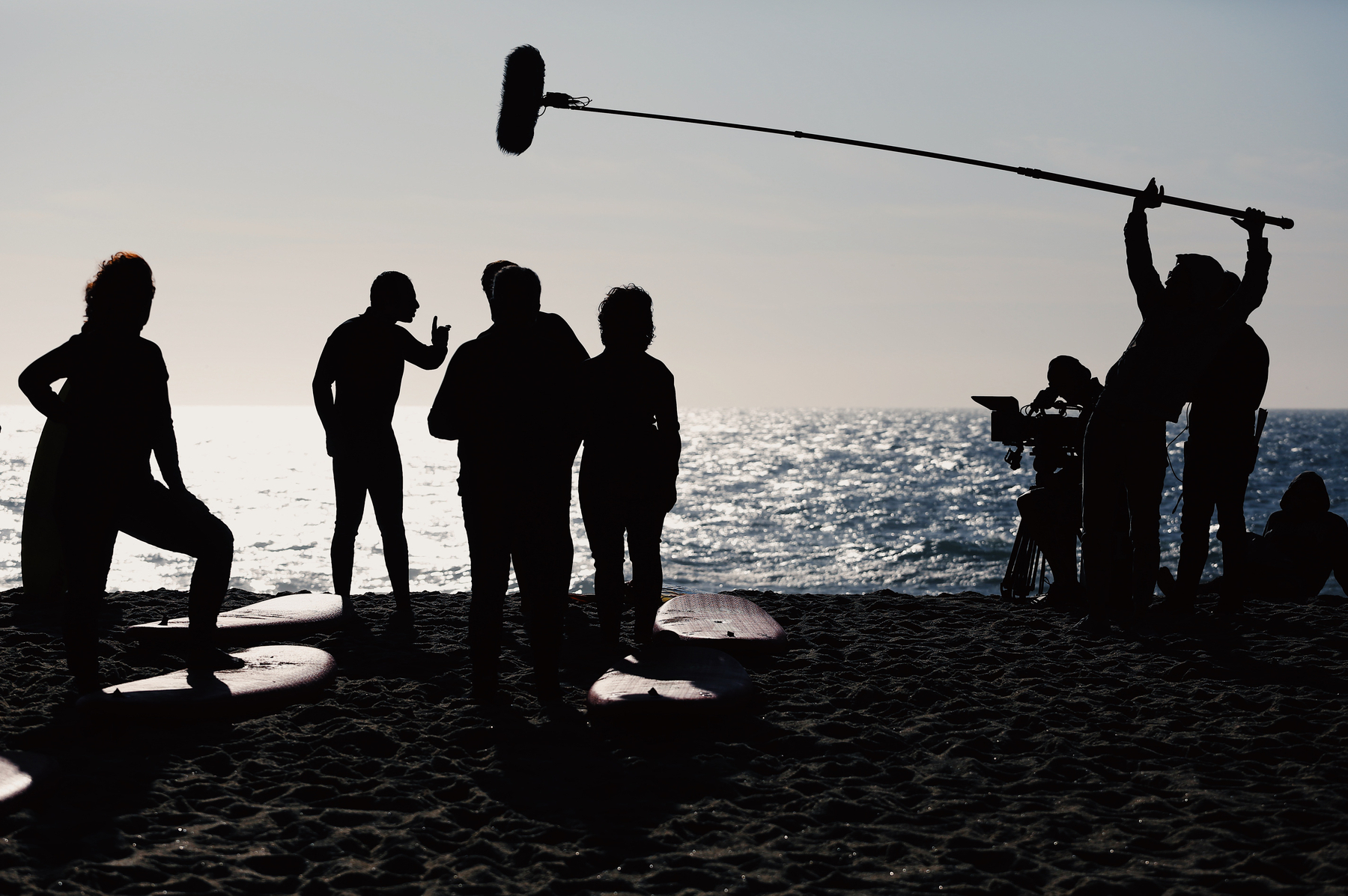 A film crew shoots a scene at the beach. We can see some surf boards, the cast on the left, crew on the right, and a huge boom microphone over them. 
