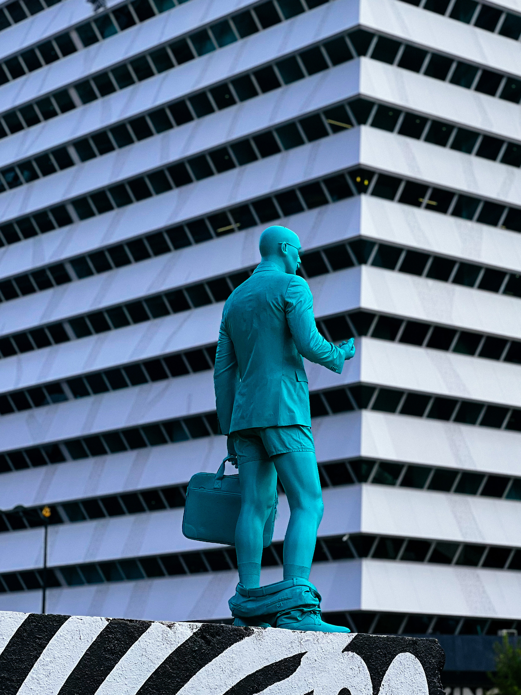 A street art piece, a green sculpture of a man holding a laptop briefcase, with his pants down. 