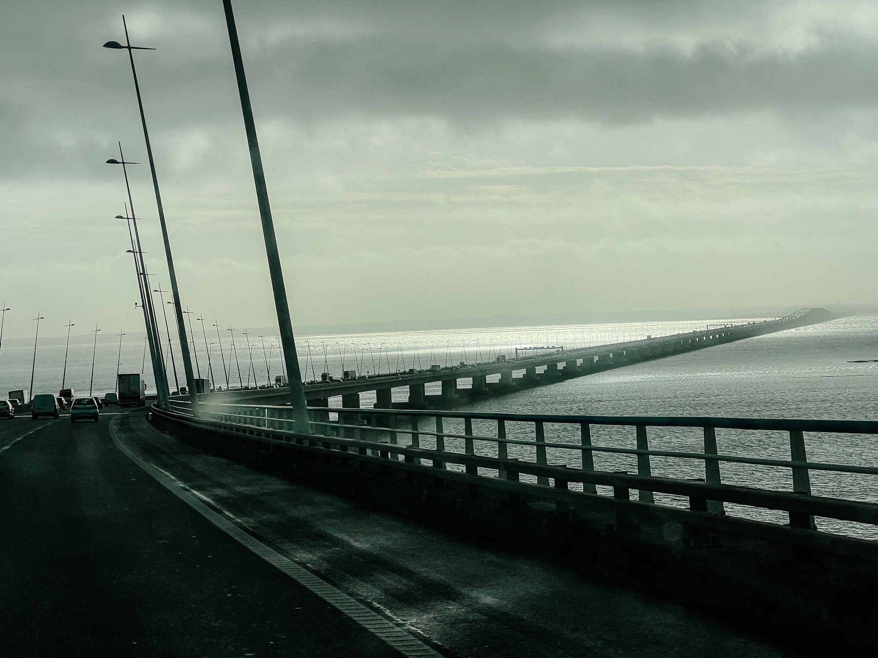 Driving on a very long bridge, we can see the wide river in front of us, and a stormy sky