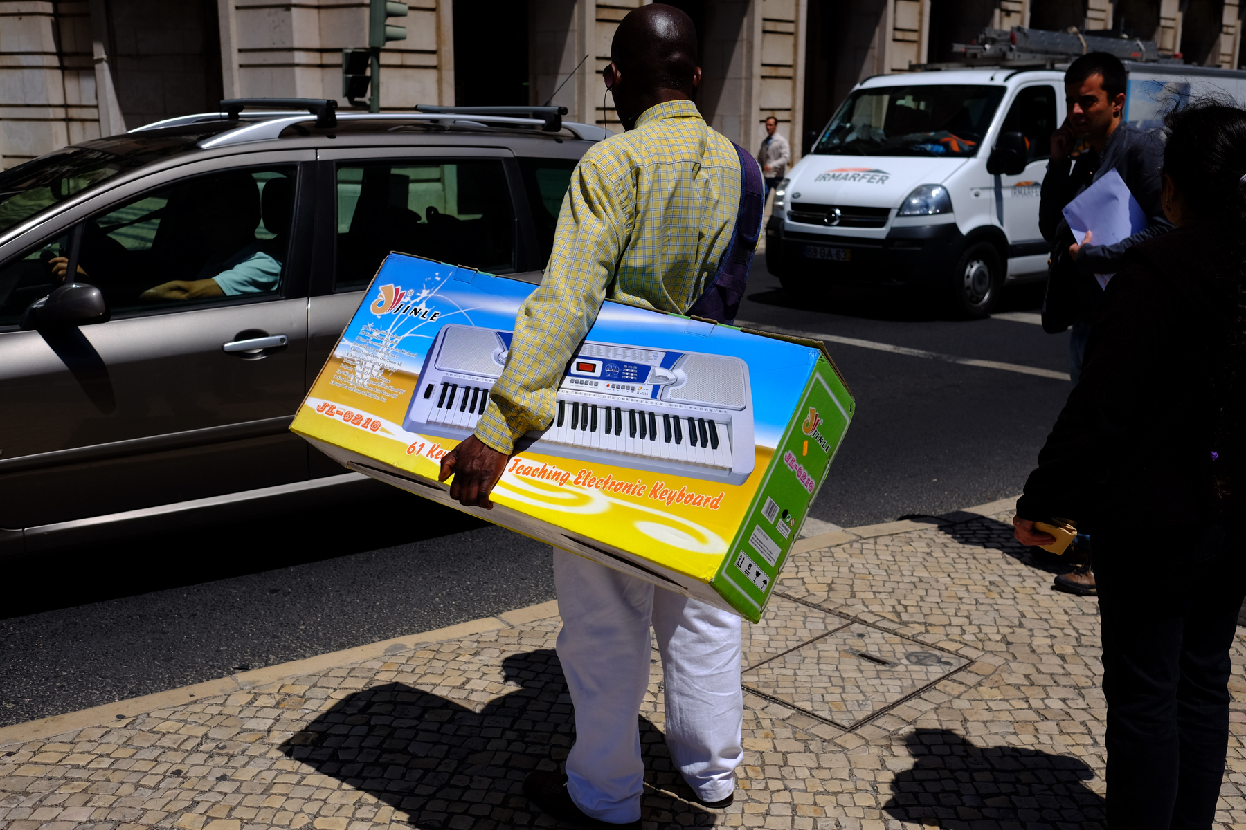 A man carrying a big box with a photo of a keyboard waits to cross a busy street. 