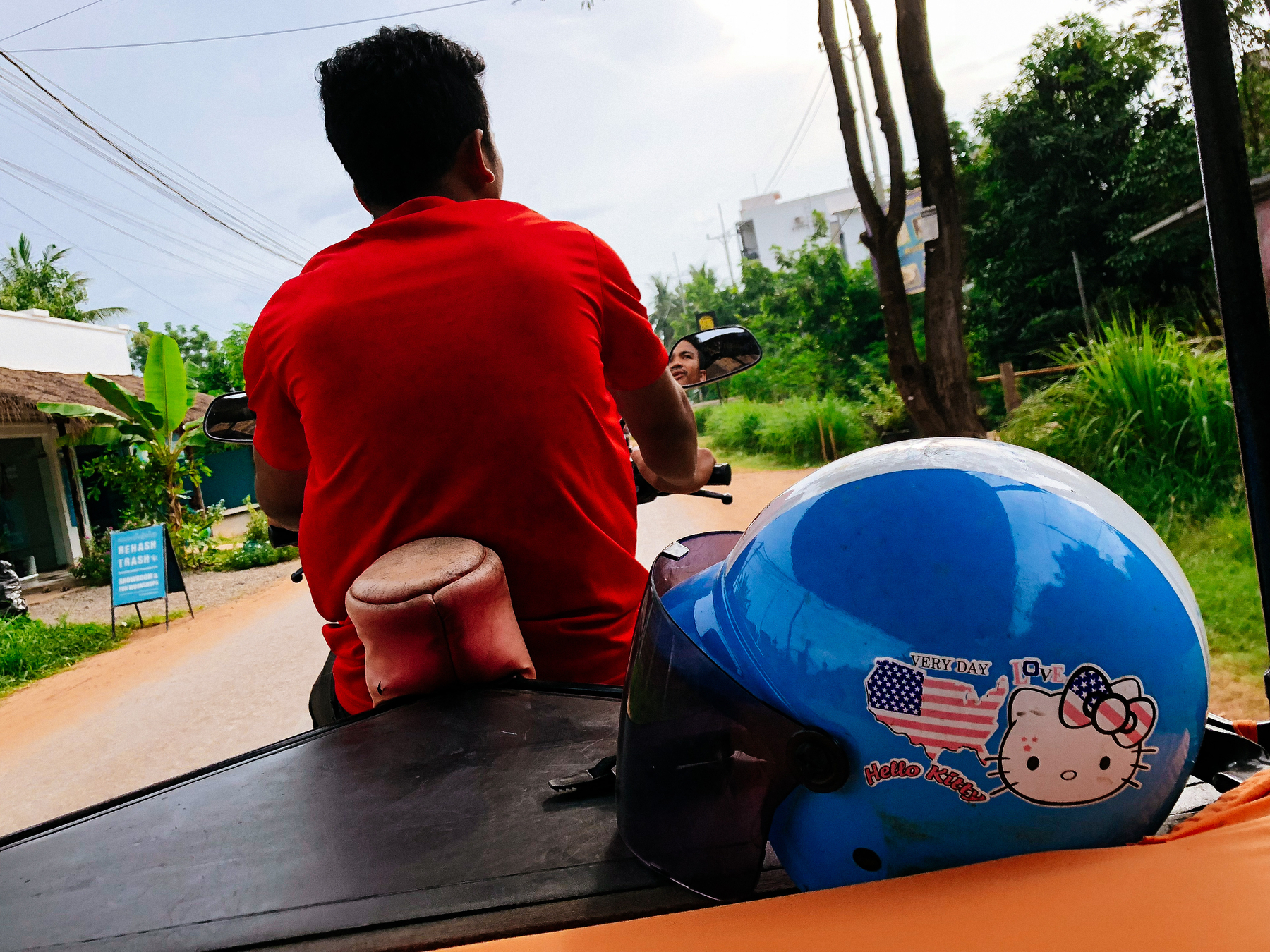 A tuk tuk driver with his back to us, and a blue helmet in the foreground, the word “very say hello Kitty love” written on it 
