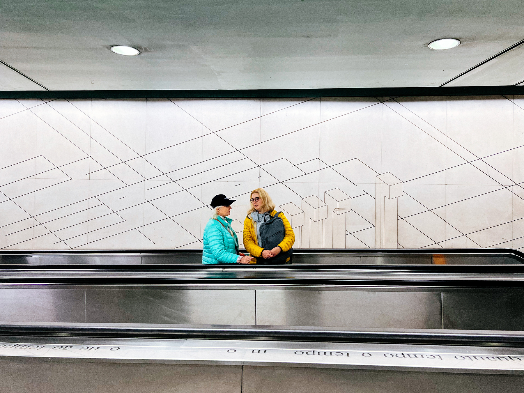 Two women ride an escalator. One in bright yellow jacket, the other light blue. 