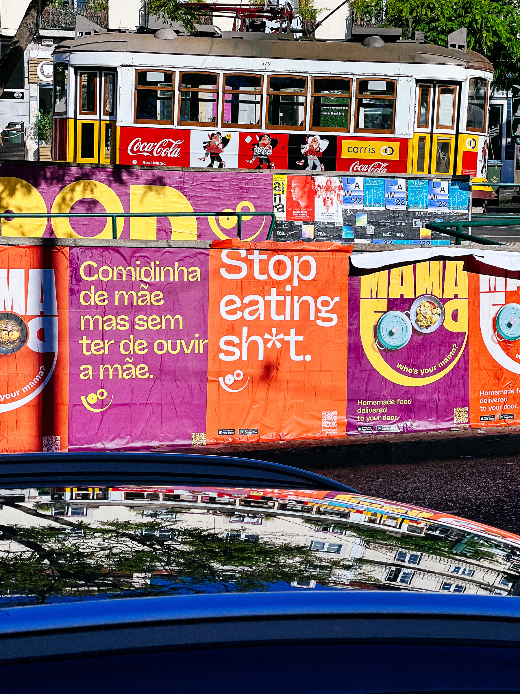A tram in the background, with posters on a wall in the foreground. One says “stop eating sh*t”