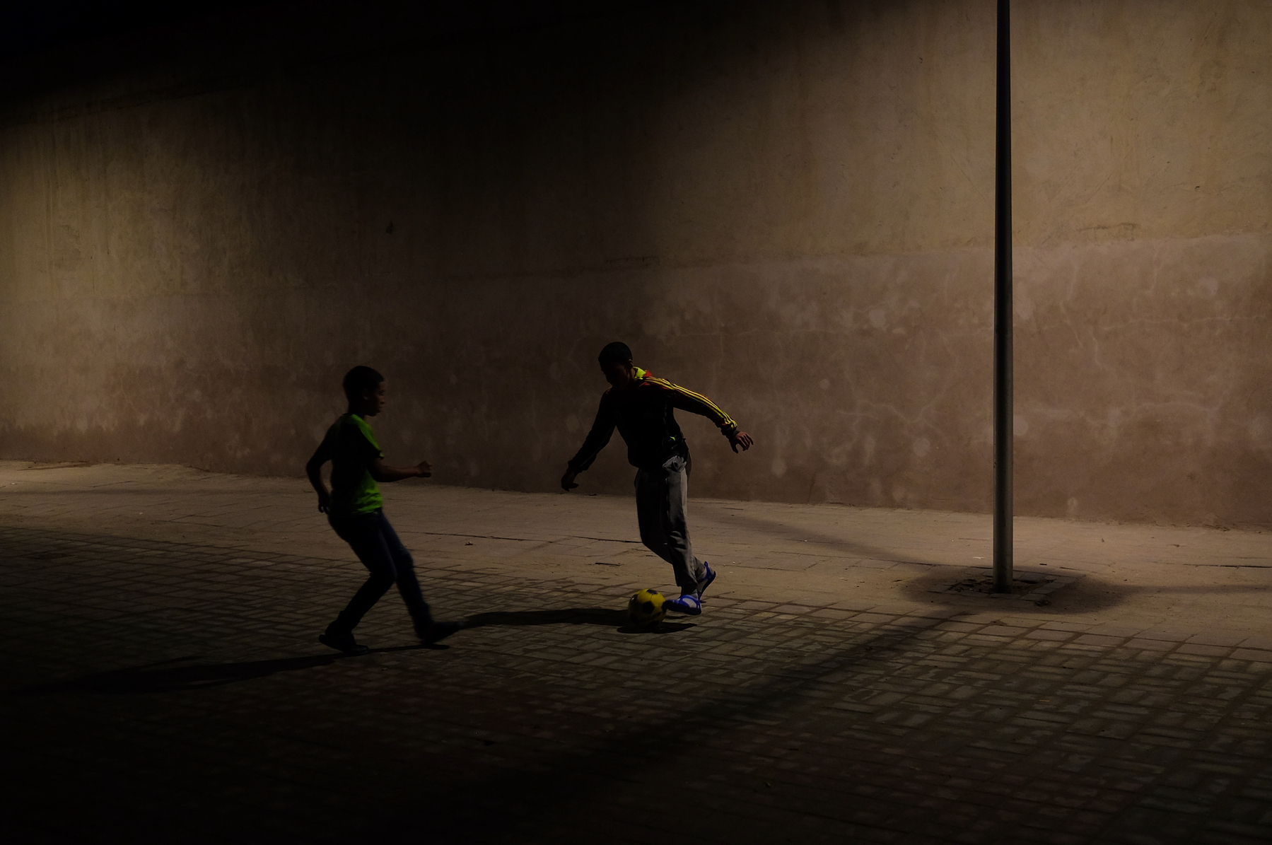 Two kids play football into the night, light from a very feeble lamp coming from above. 