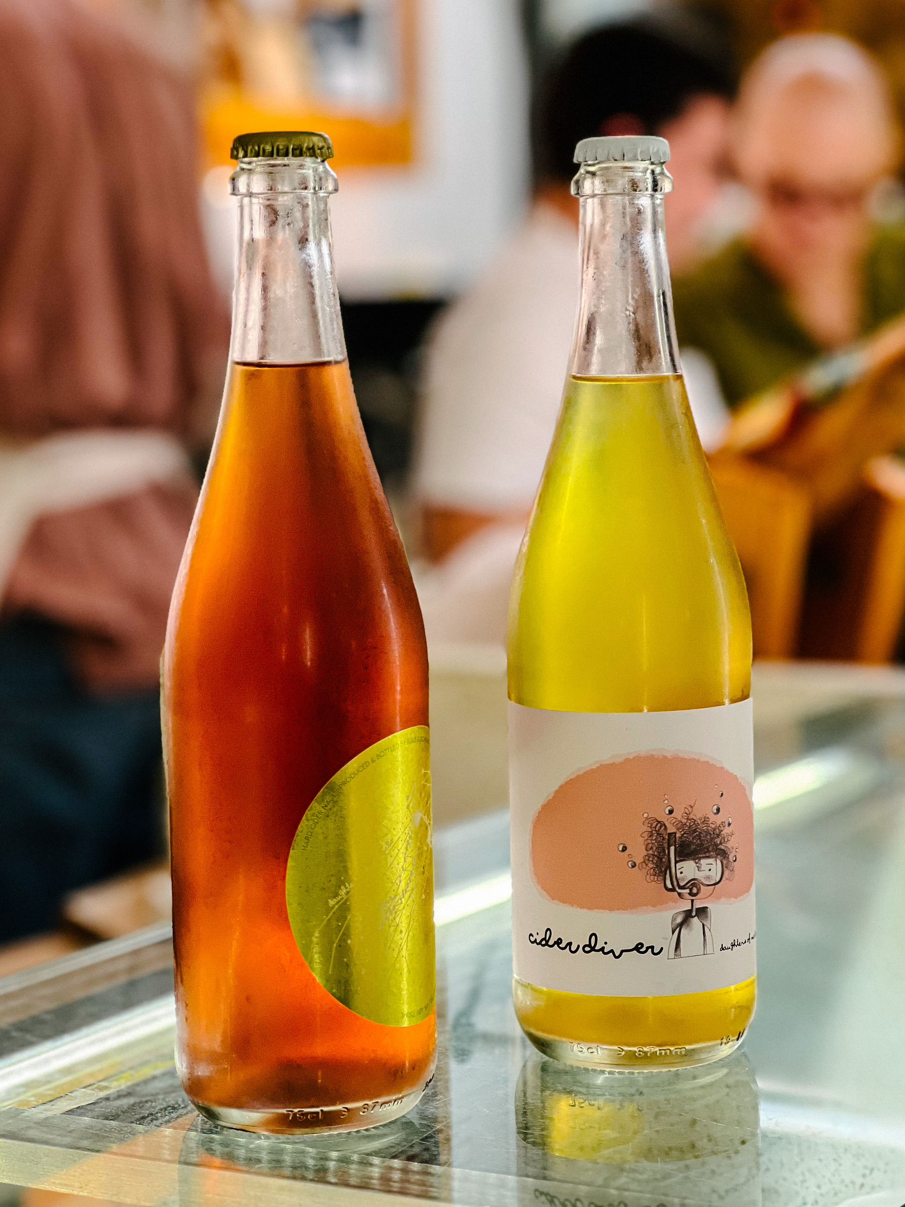 Two bottles, one filled with yellow liquid and “Cider Diver” written on the label, the other orange. People in the back. 