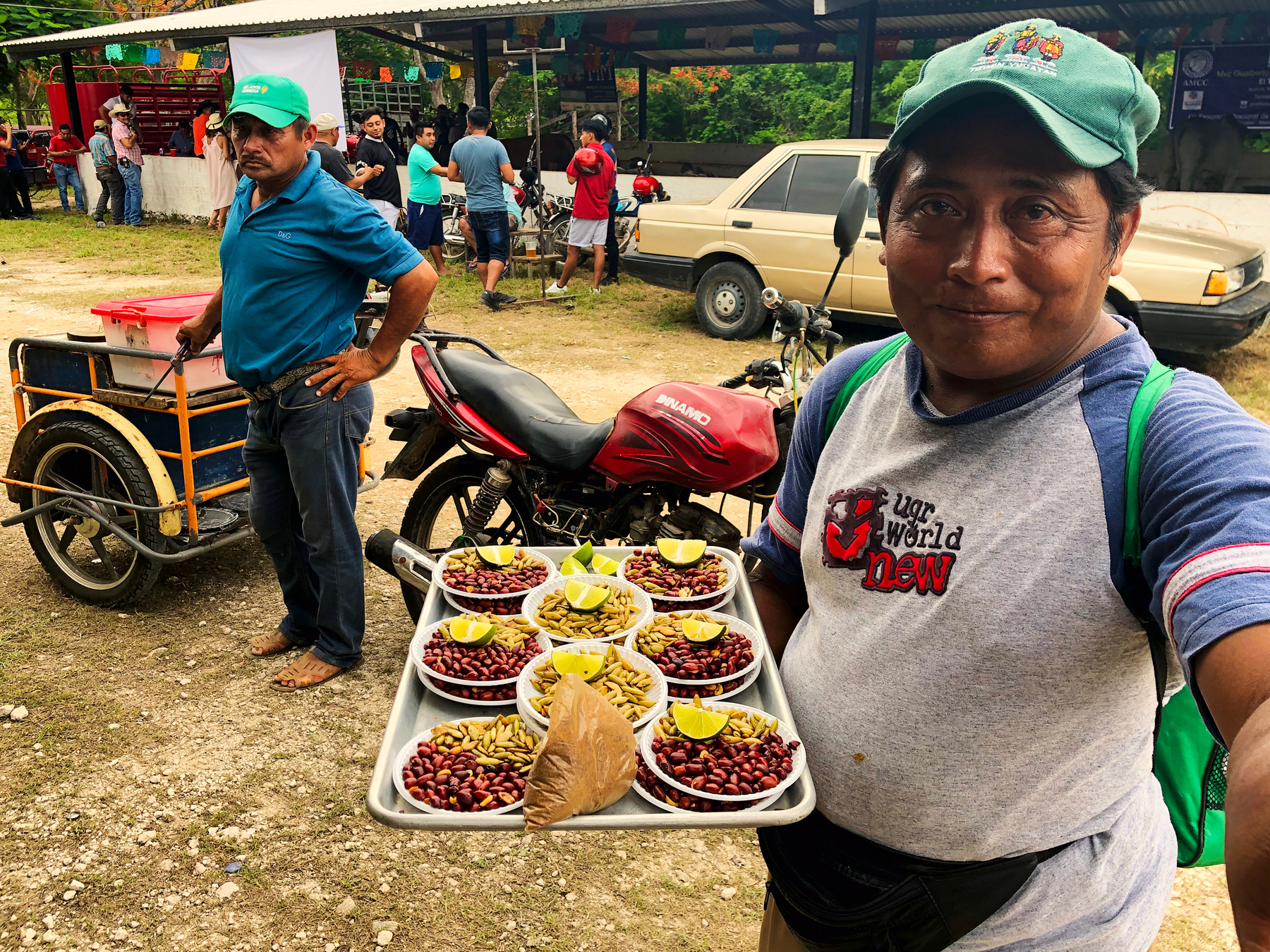 A man shows dishes with assorted nuts and lime slices. In the back other people, and a car. 