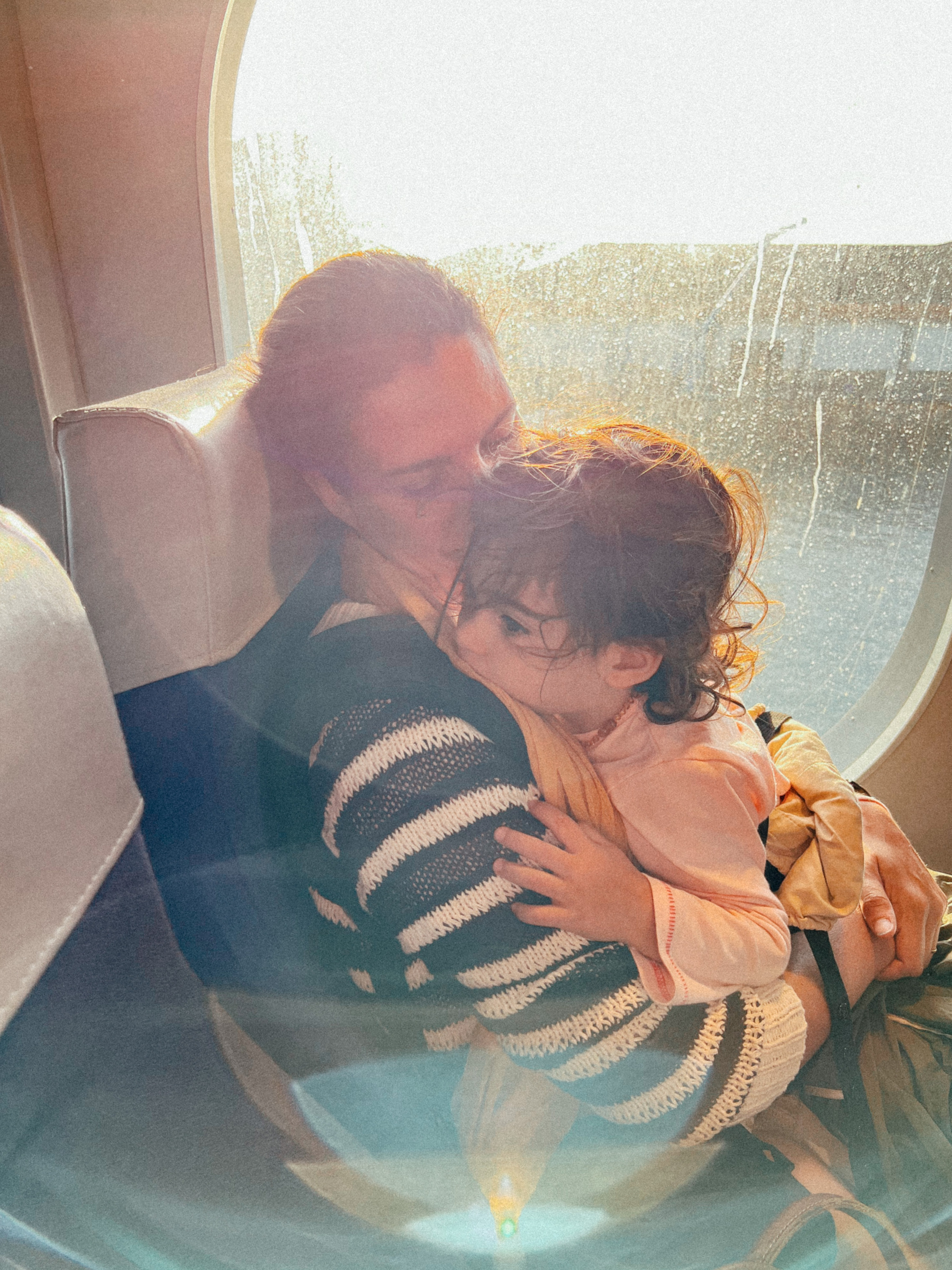 A mom and a toddler sit close to a window, hugging. 
