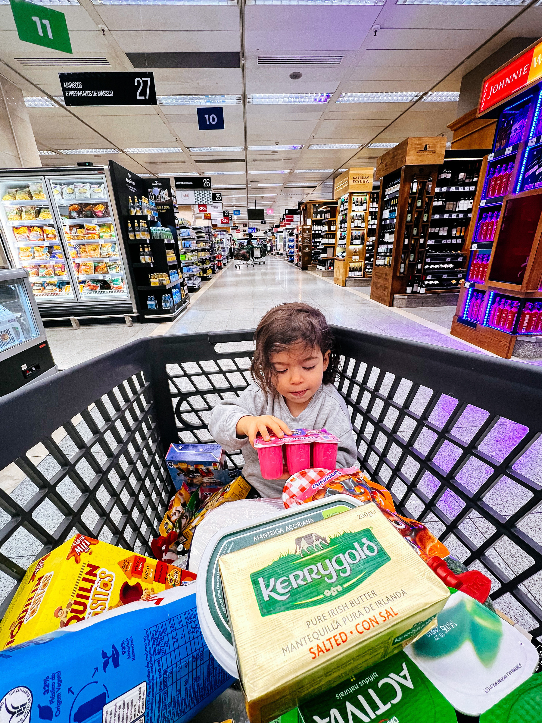 Toddler rides in a supermarket cart filled with grocery items.