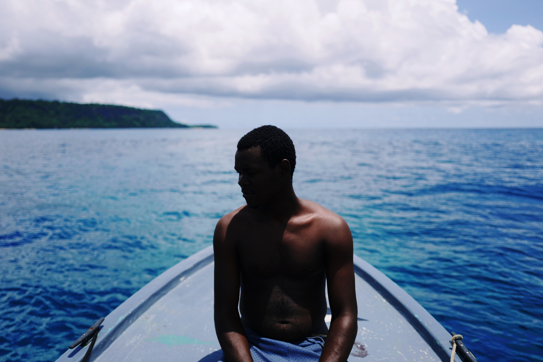 a man sits on a boat, pensive, with blue water and sky all around him.
