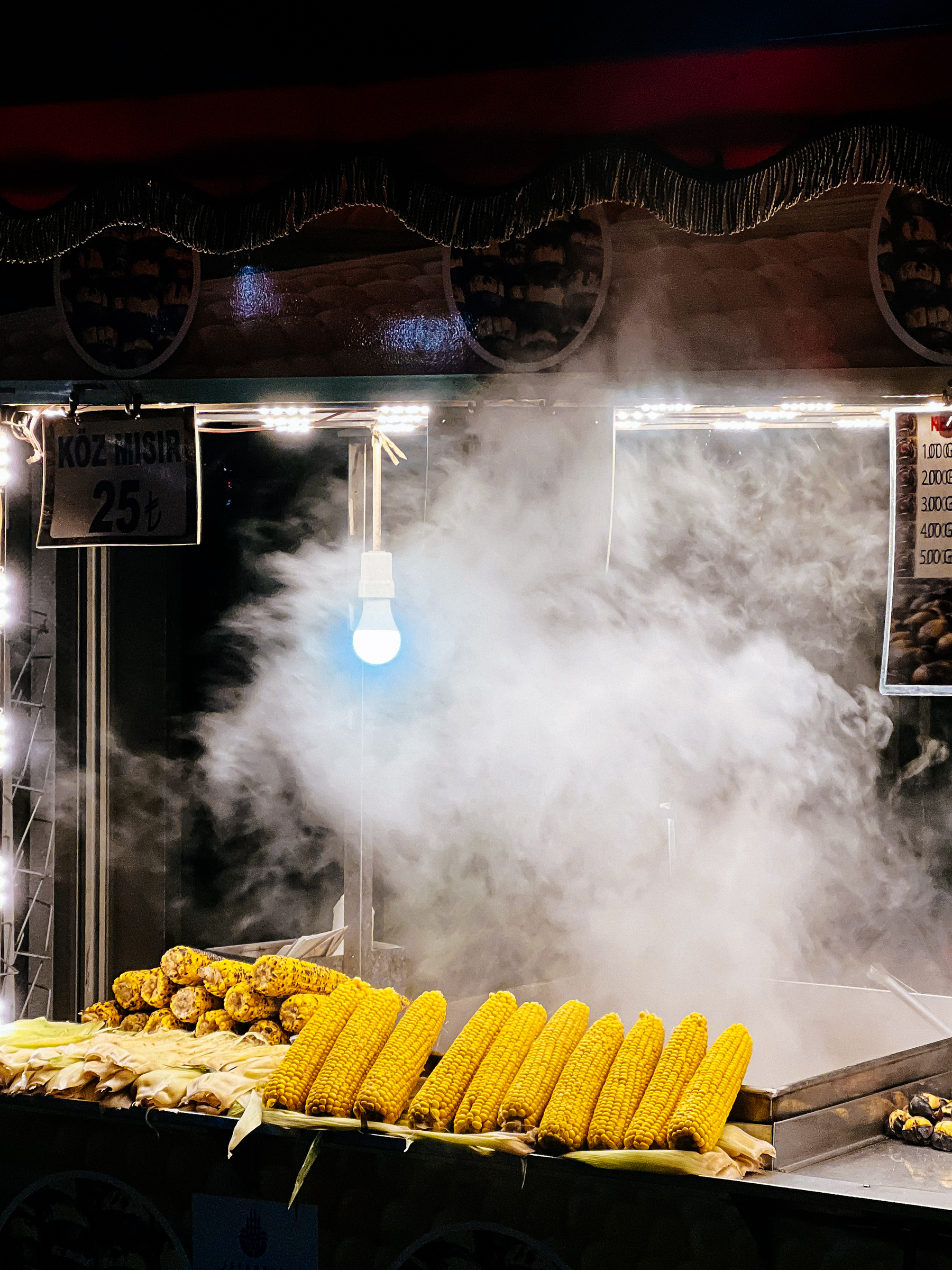 Detail of a roasted corn stall, smoke coming out of it, and plenty of corn, ready to eat. 