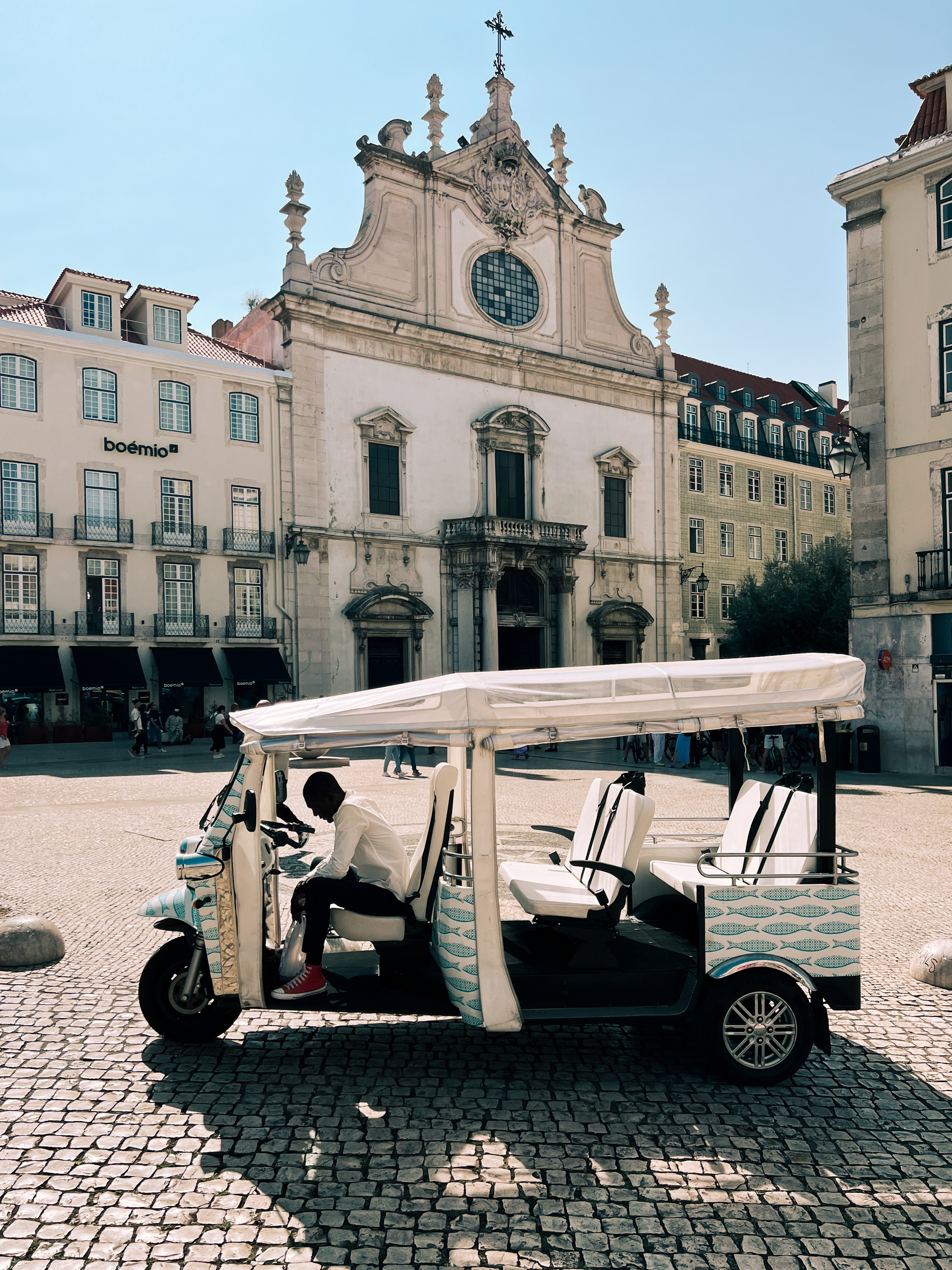 A tuk tuk waits for a customer, in front of a church. The driver is sitting in the driver’s seat. 