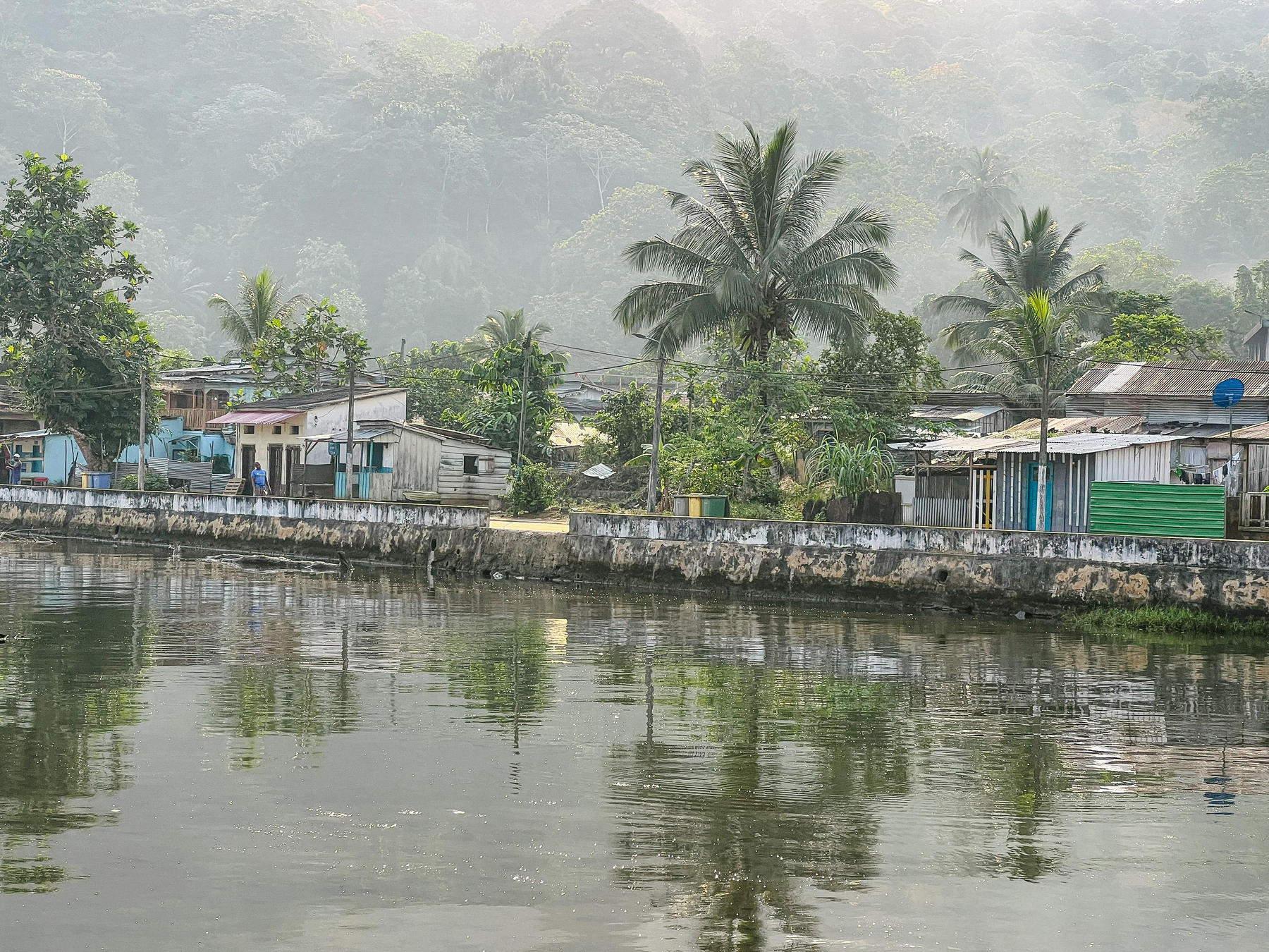 Some houses are visible, with a river in front of us, and rainforest in the background 
