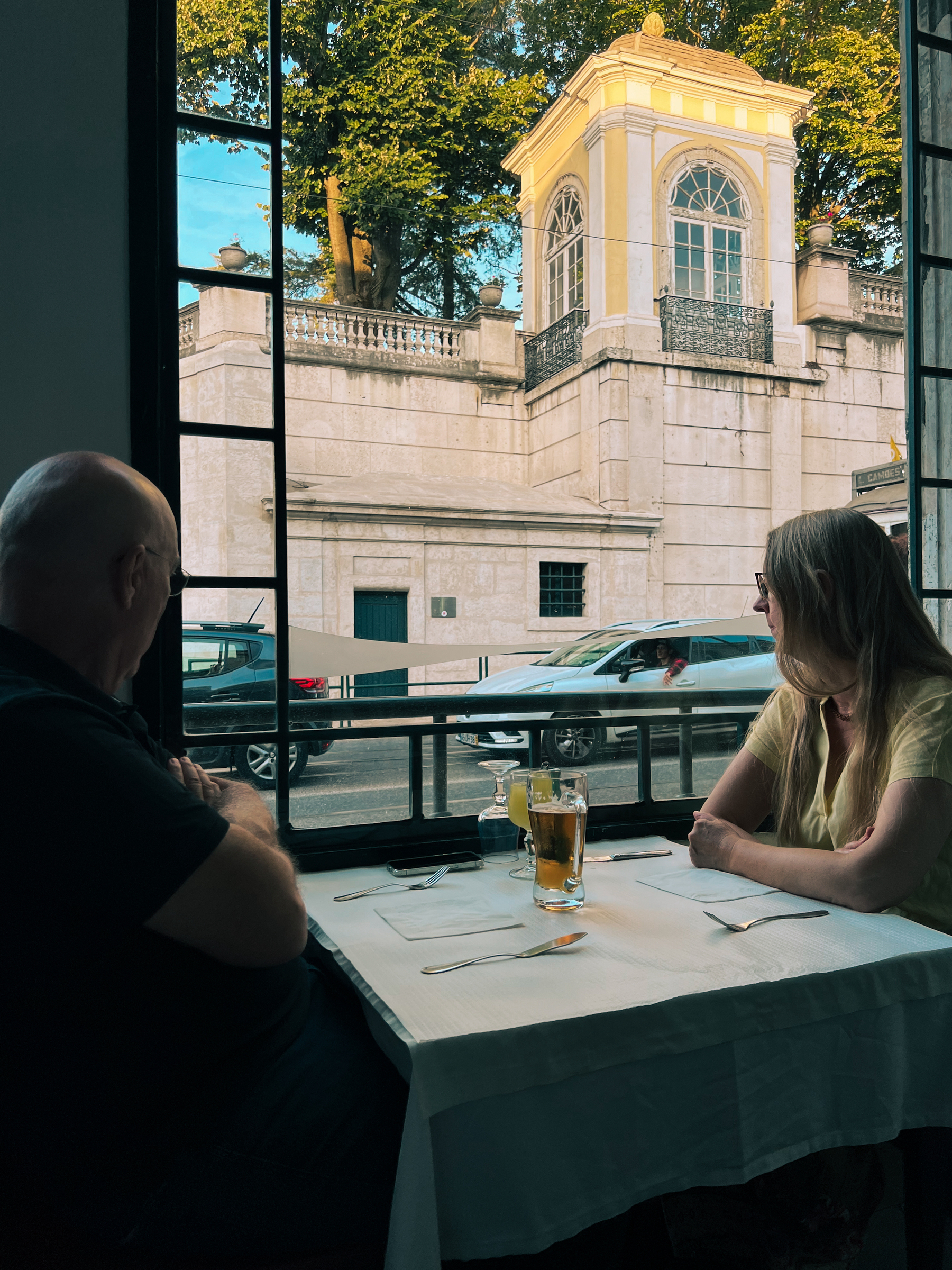 Two people look out the window at a restaurant, a beer and a juice on the table. Outside some cars drive by a big wall with a small “tower” (is there a word for this type of thing?)