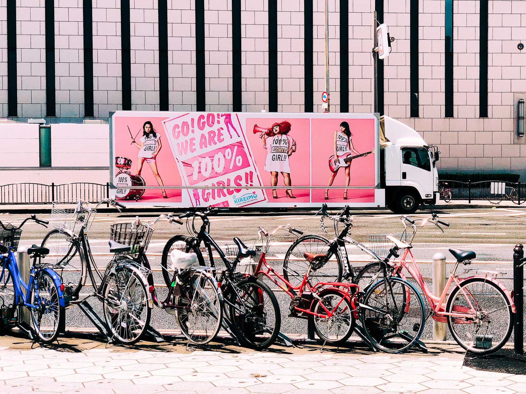 A truck with a pink ad for “Go! Go! We are 100% Girls!”, with a row of bicycles in the foreground. 