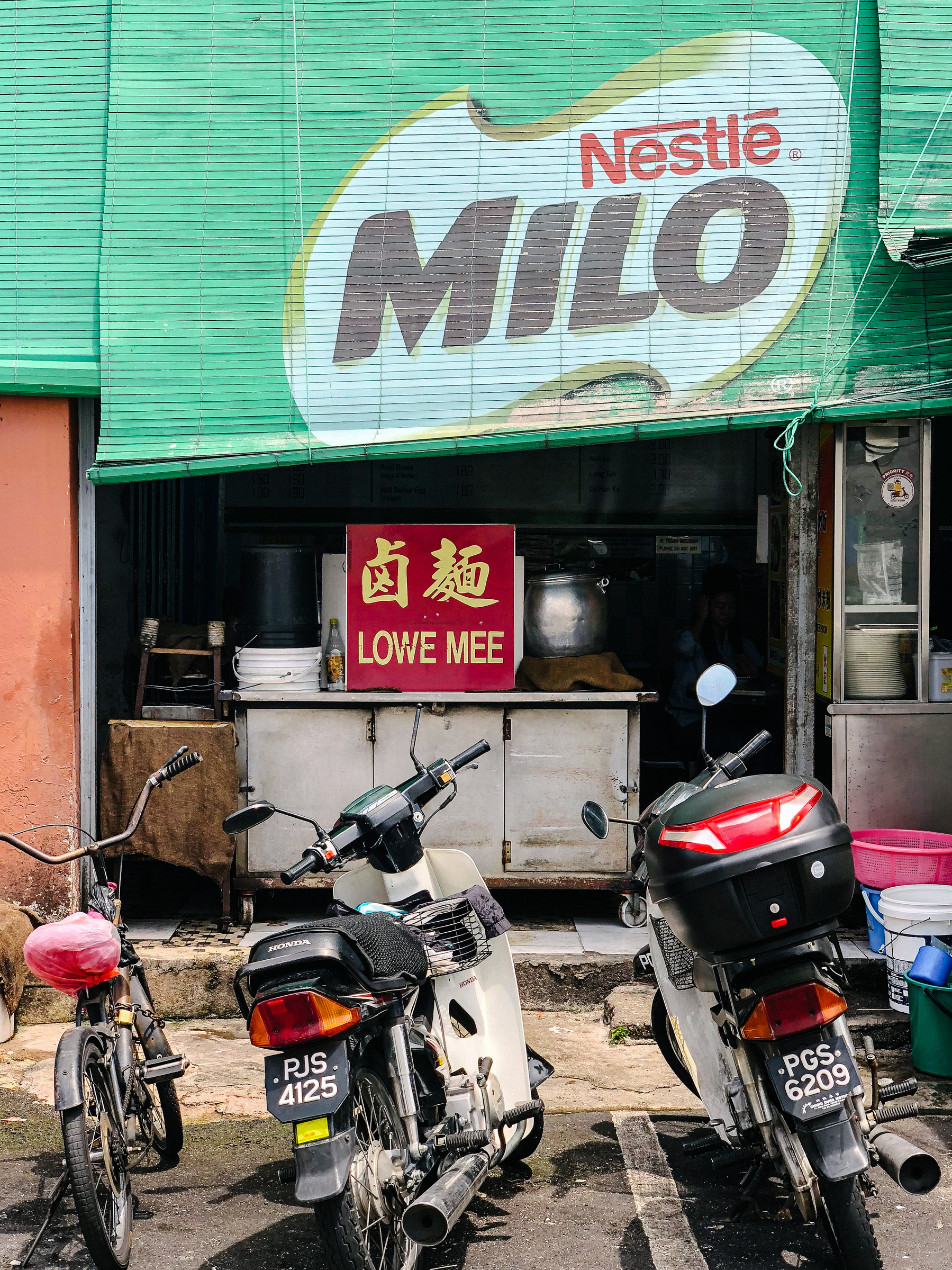 A store front with a huge ad for Milo, parked motorcycles, and a sign that reads “lowe mee”. 