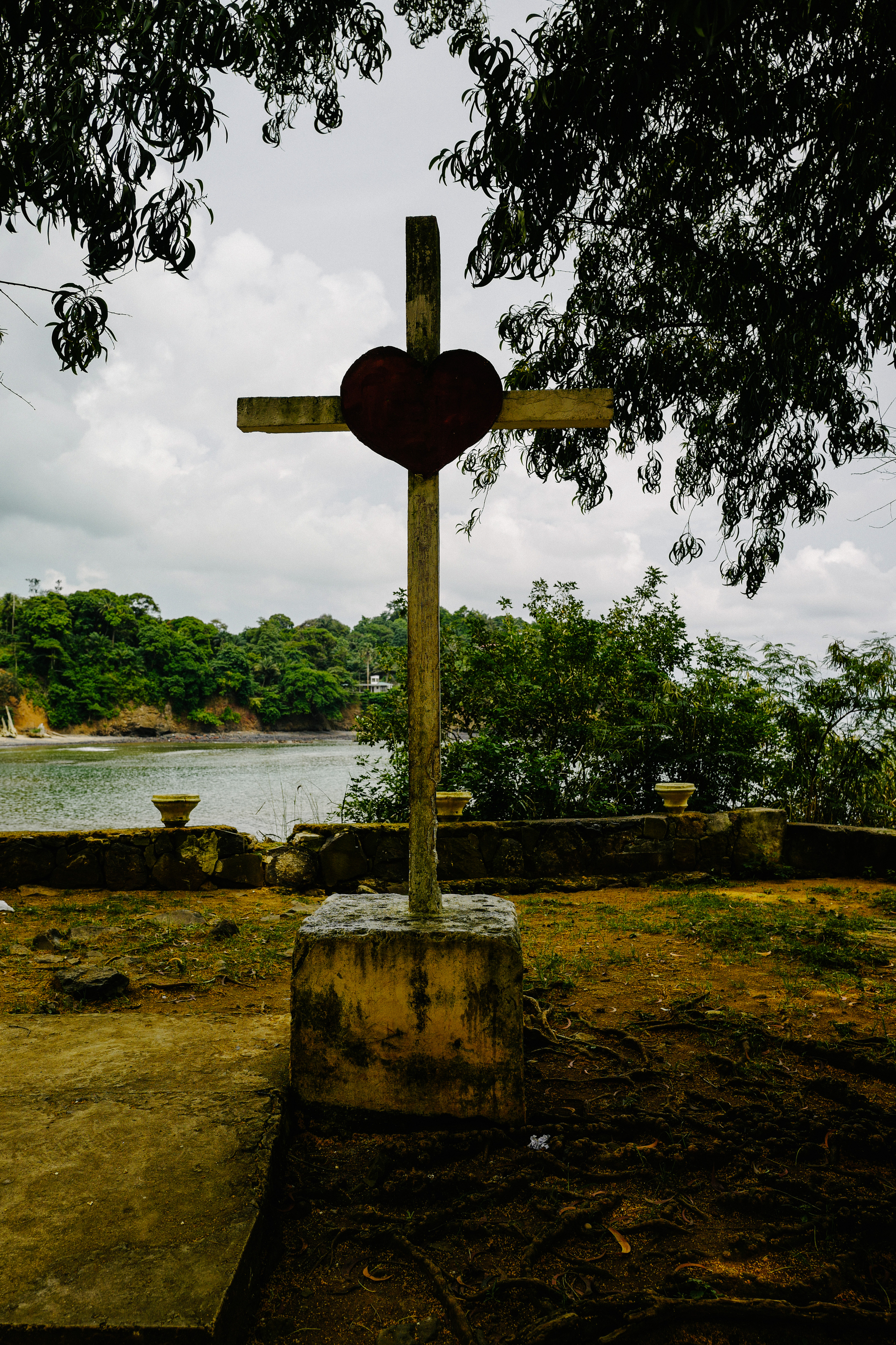 A wooden cross with a heart instead of an image of Jesus. It stands under a tree, facing the sea. 