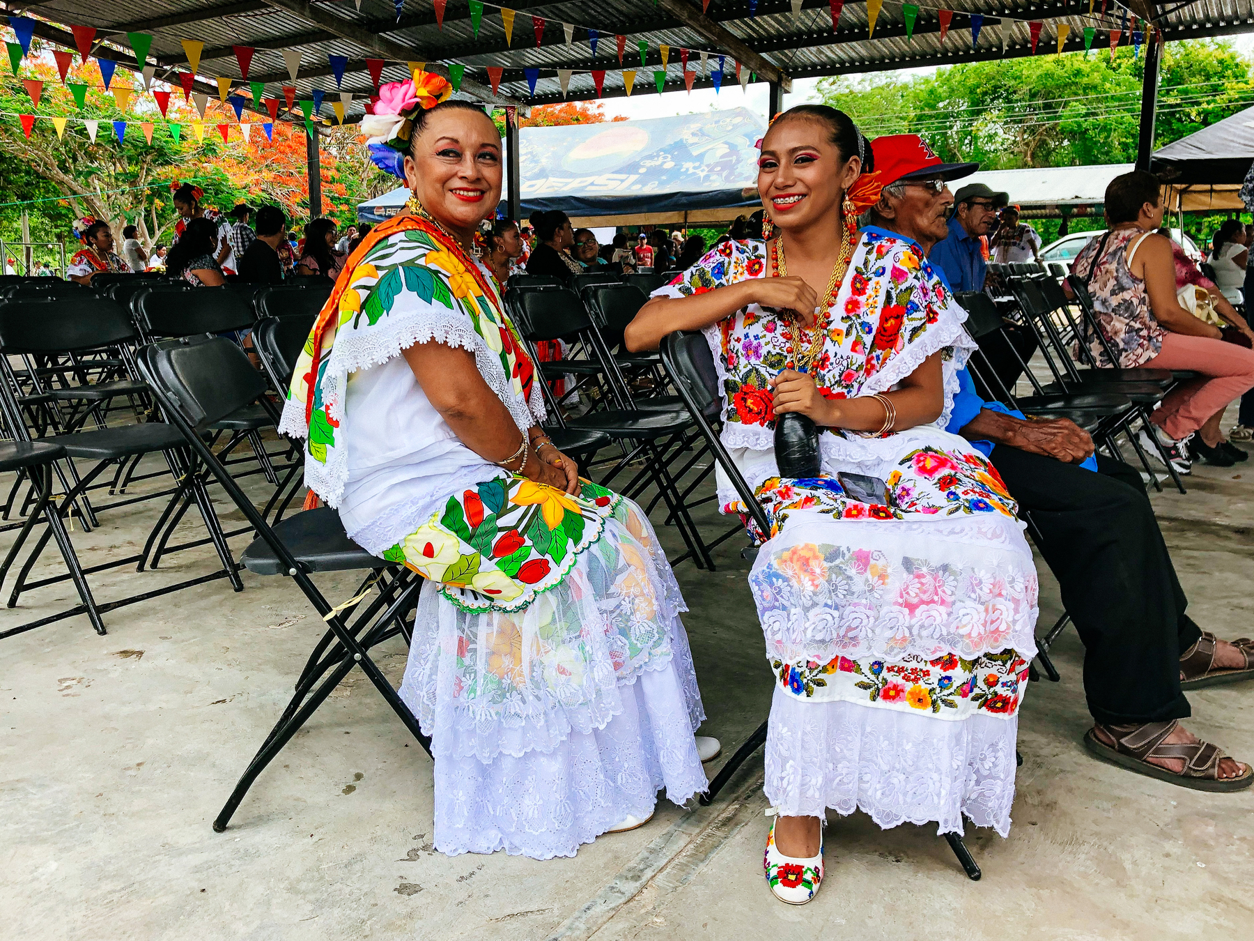 Two women in traditional Mexican dresses sit in chairs, in what looks like a party. 