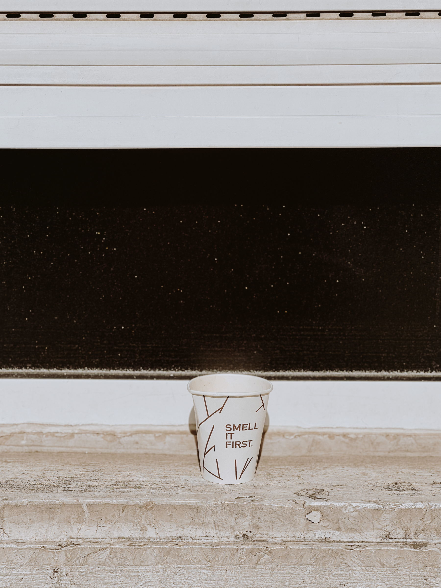 A paper coffee cup rests on a window sill. “Smell it first”. 