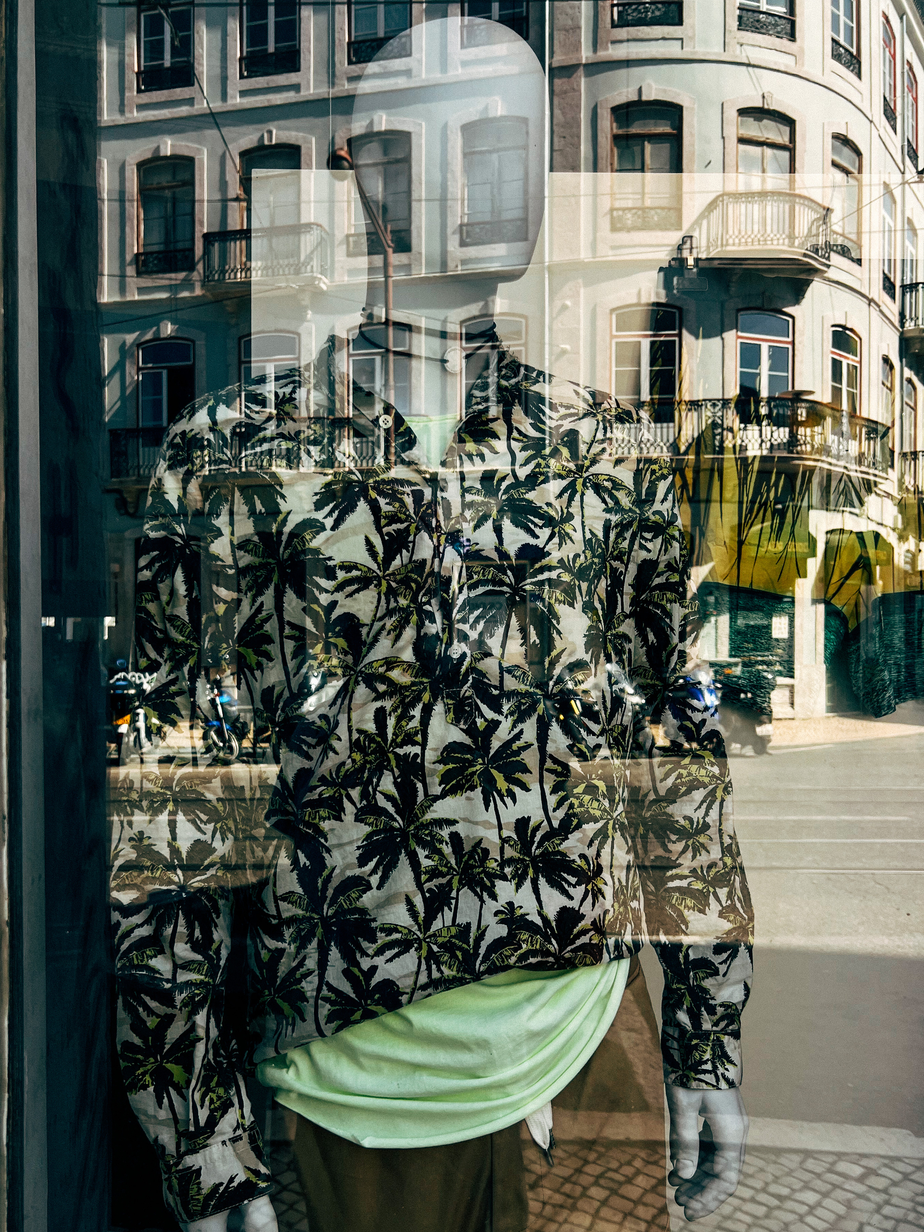A mannequin wearing an awesome shirt with palm trees on it, and a building reflected on the shop window. 