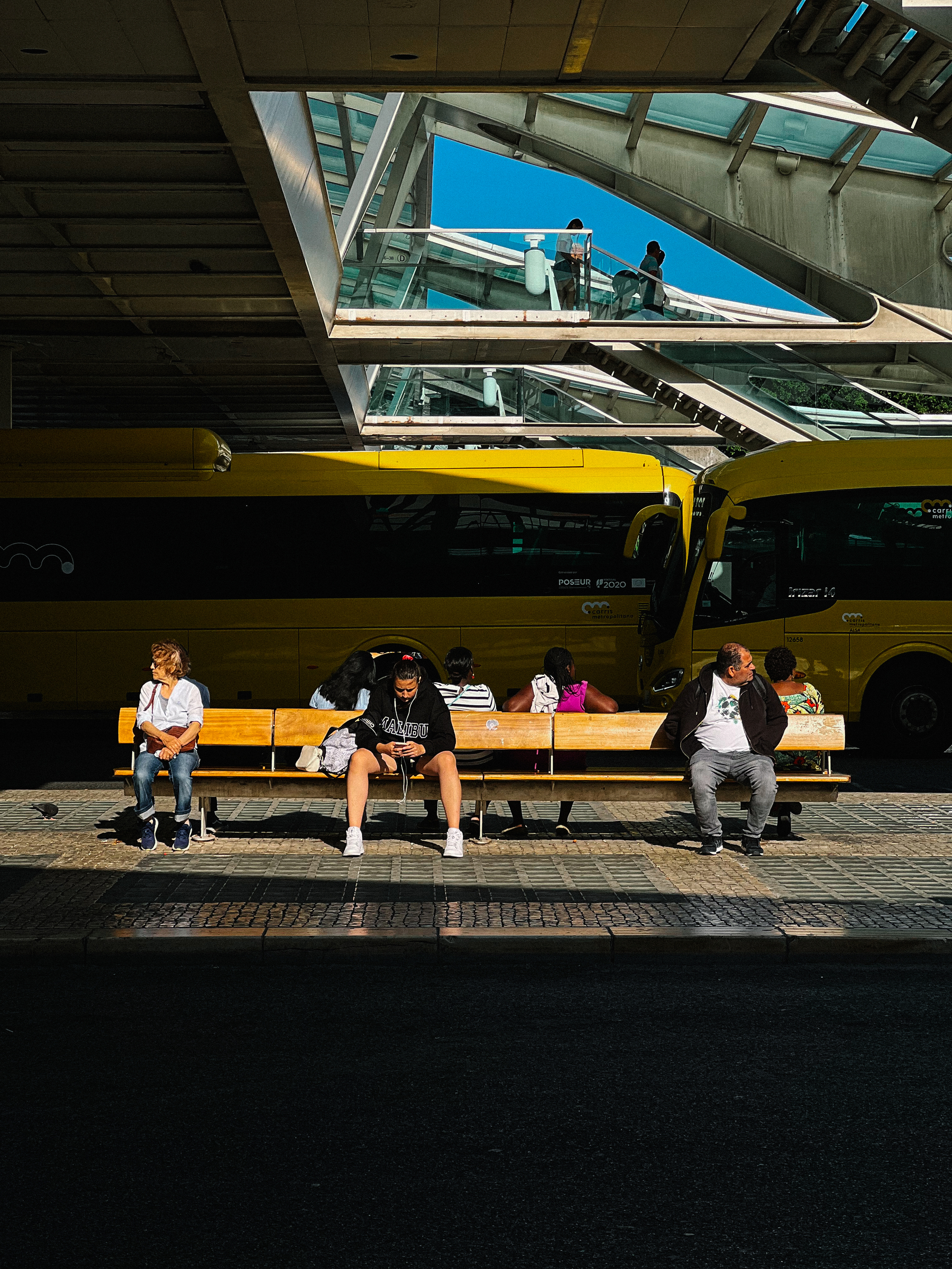 People sitting, waiting for a bus. Buses in the background. 