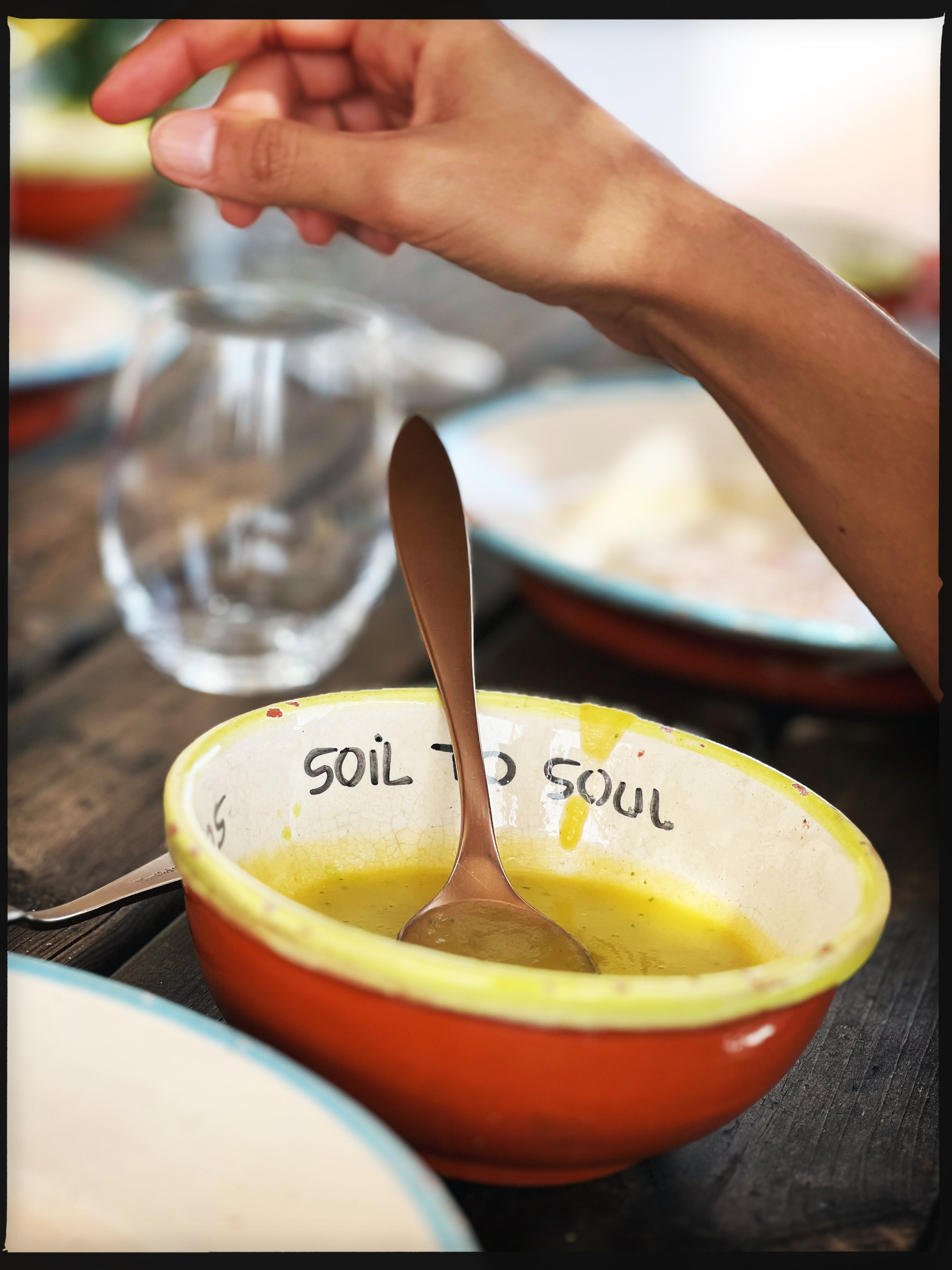 A soup bowl with “Soil to Soul” written on the inside. There’s a hand, and a spoon. 