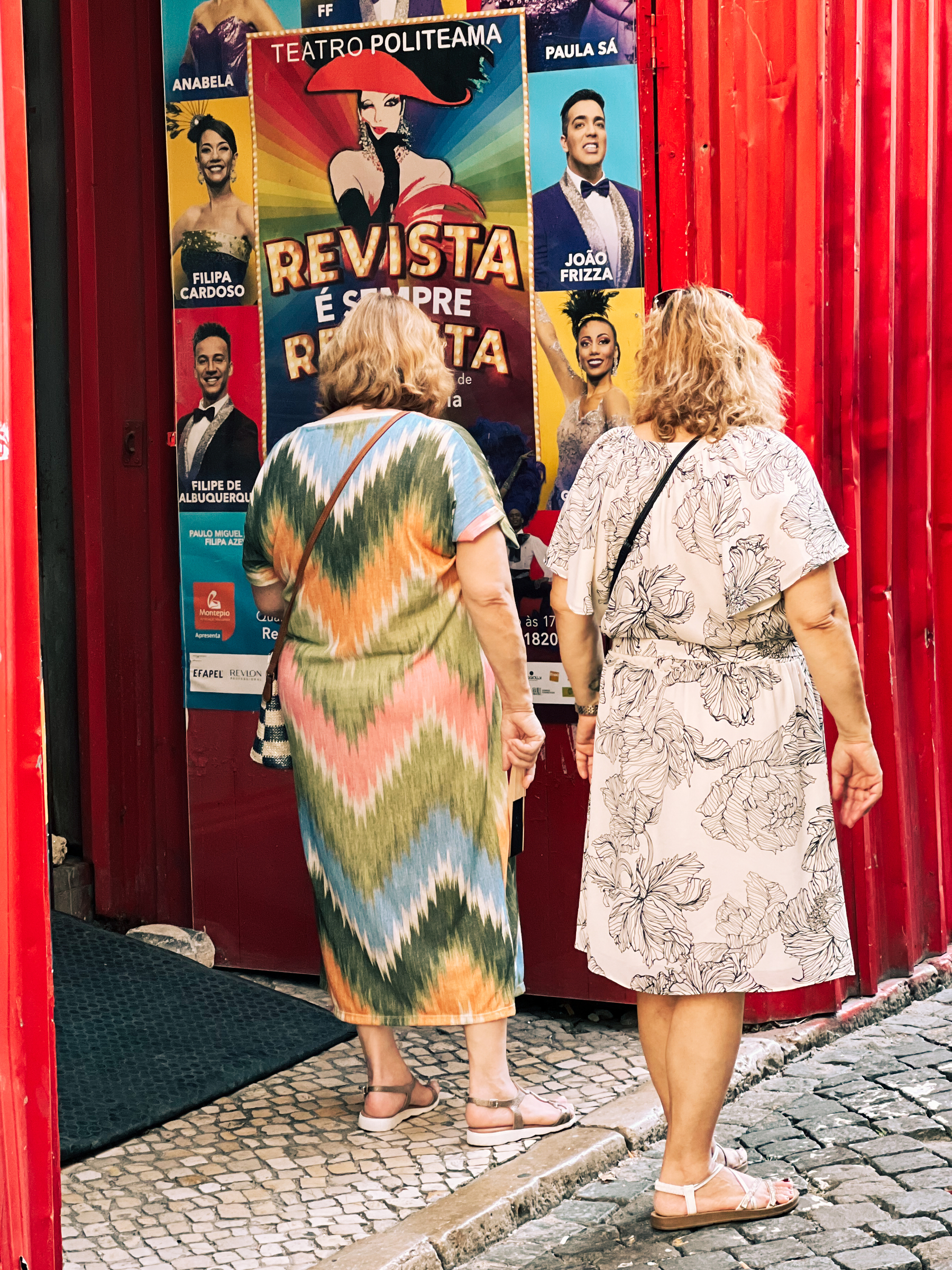 Two women look at a show’s poster on a red wall, their backs to us.