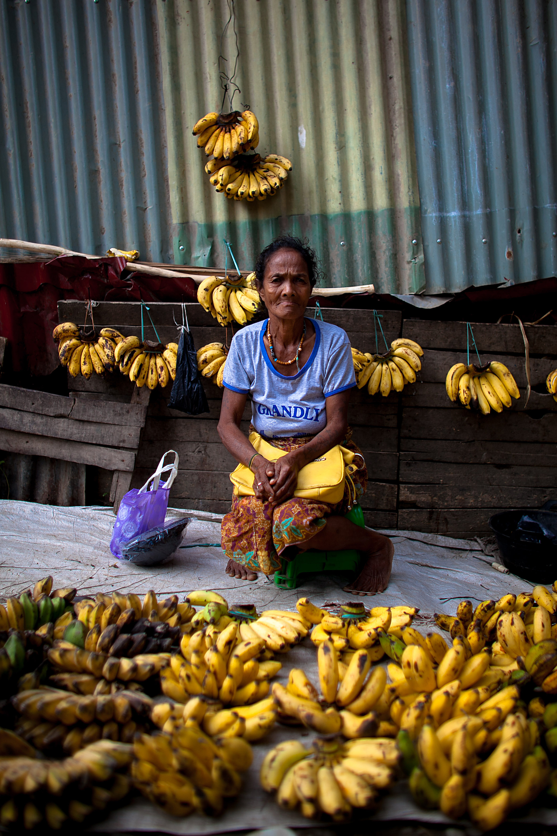 A woman sits down with bananas in front of her, and hanging from a wall behind her. She’s in the market, selling them. 