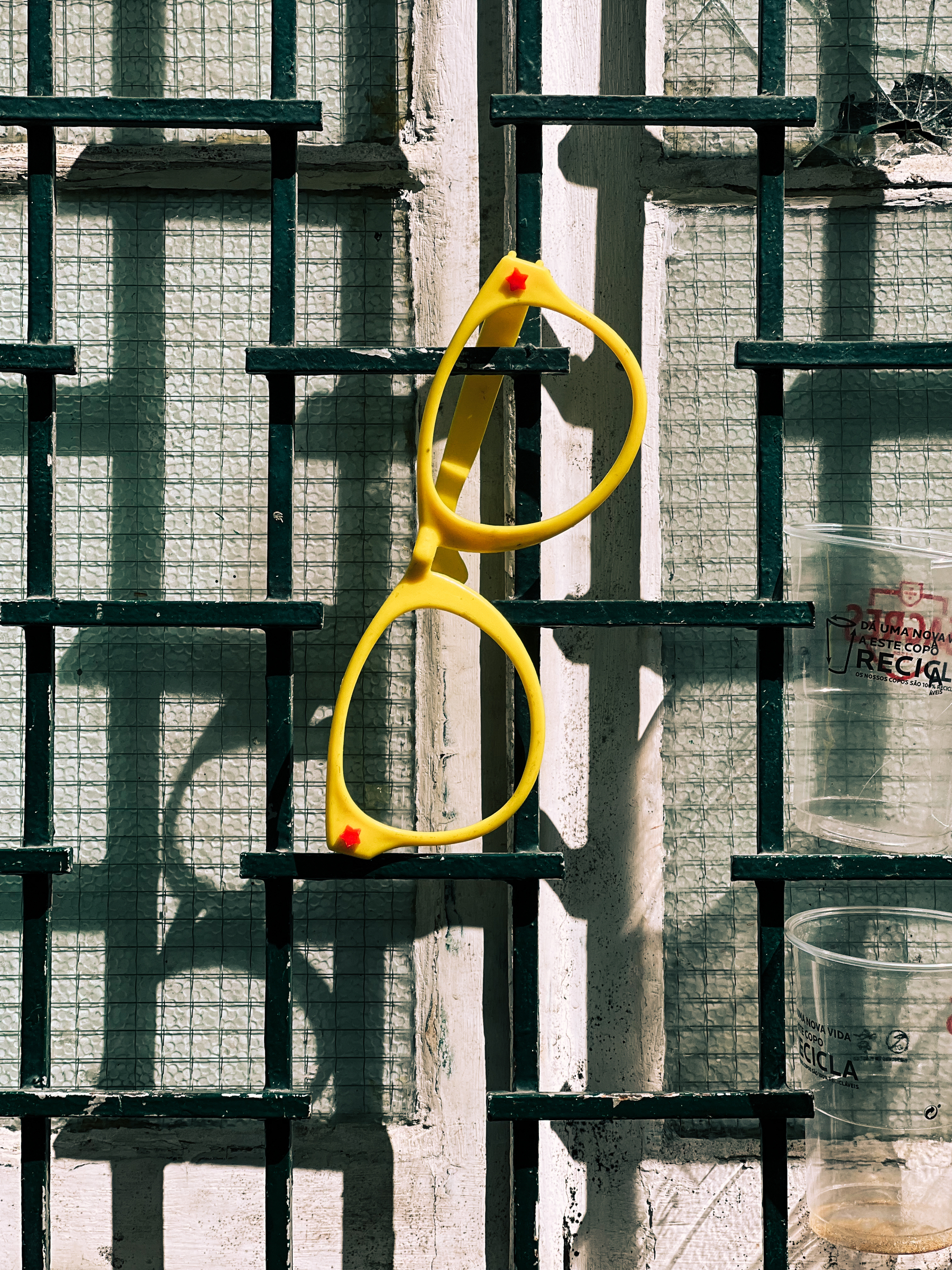 A pair of giant yellow glasses (no lenses), hangs from a window, next to discarded plastic cups. 