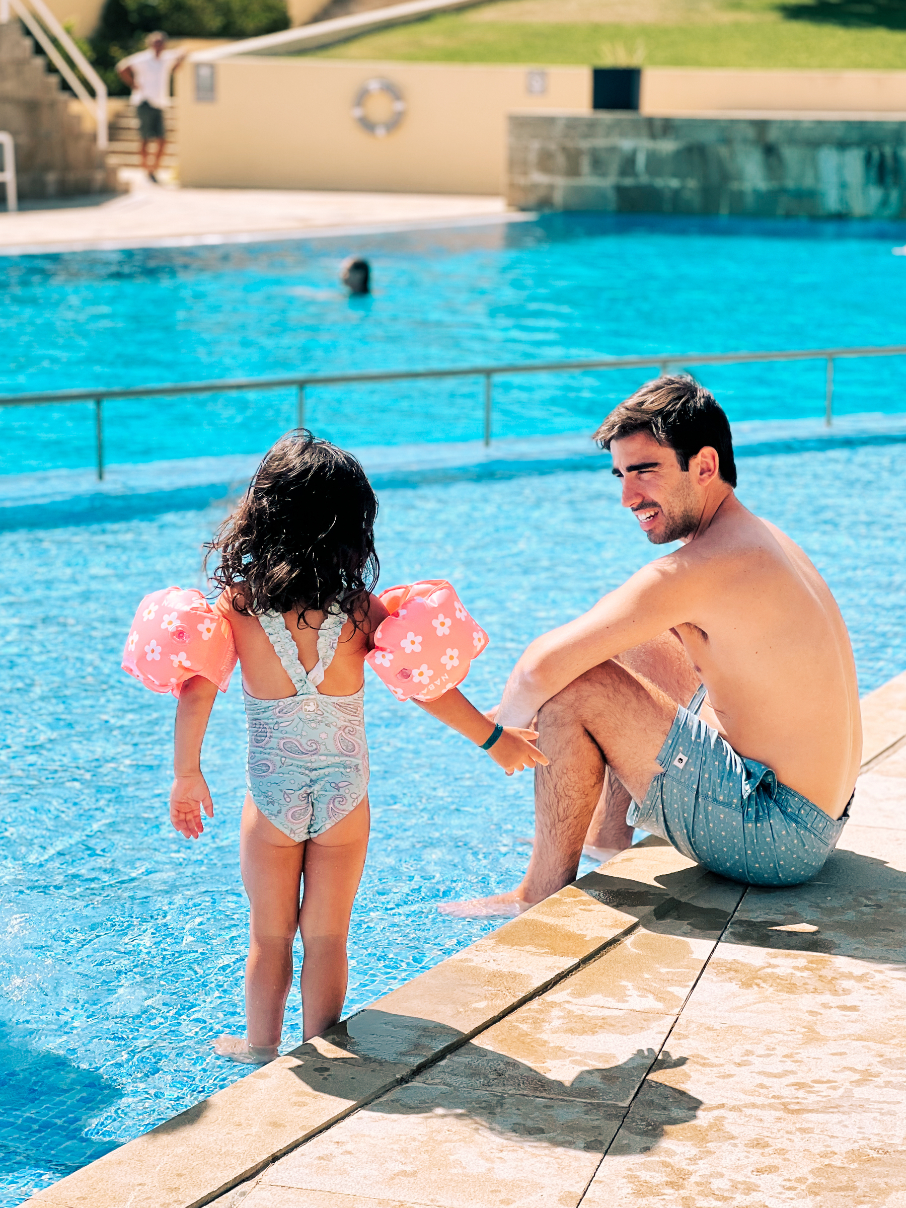 A girl standing in a pool, wearing floaters, talks to a man who’s sitting next to her. 