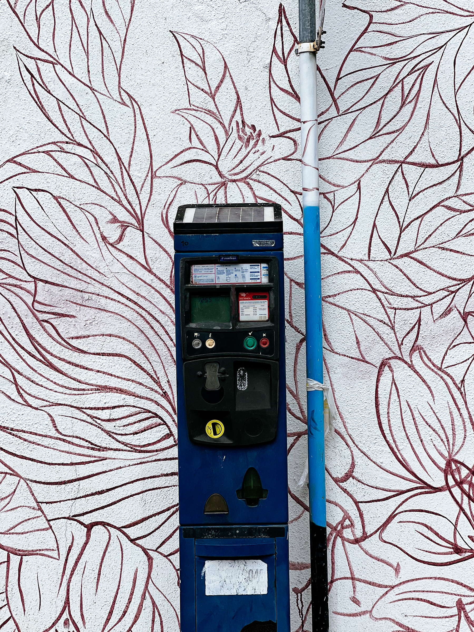 A Parking meter ticker machine stands next to a wall painted with flowers. 