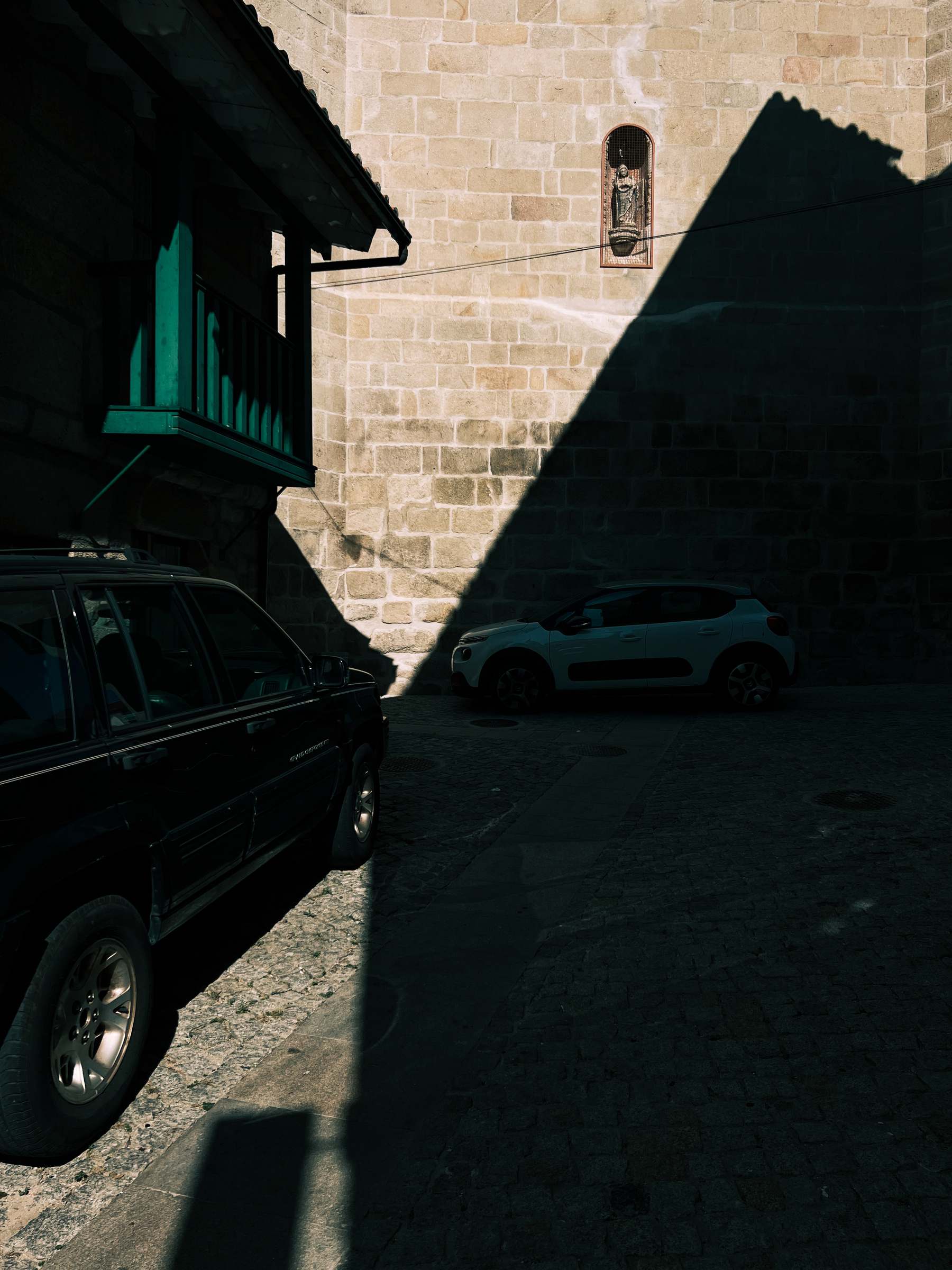 Cars parked in a alley behind a church. There’s an image of a saint, a very small one. Shadows. 