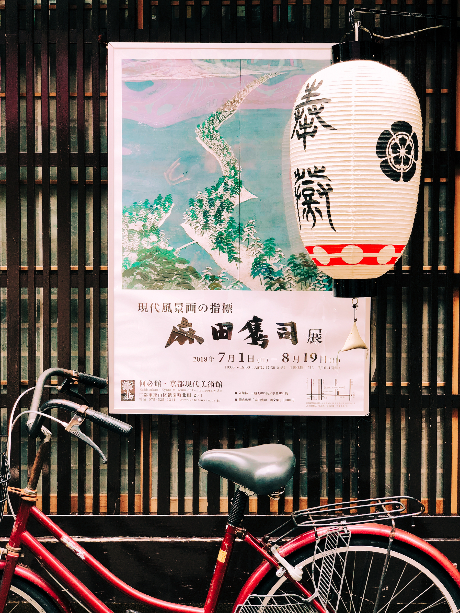 The scene is set in Japan. A bicycle is parked against a wall where a lovely poster is hanging. There’s a lamp next to it. 