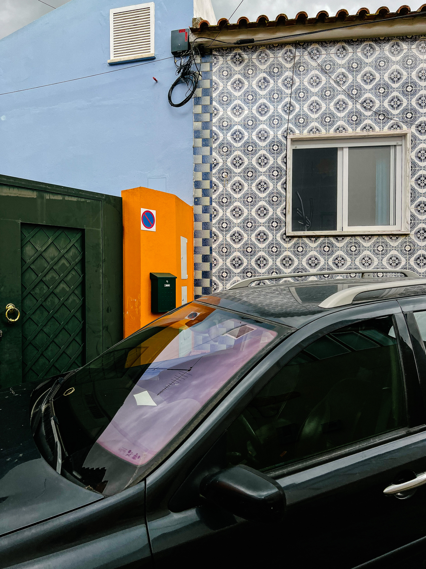A car is parked outside a crazy looking corner, with a tiled house, with a green and orange door. 