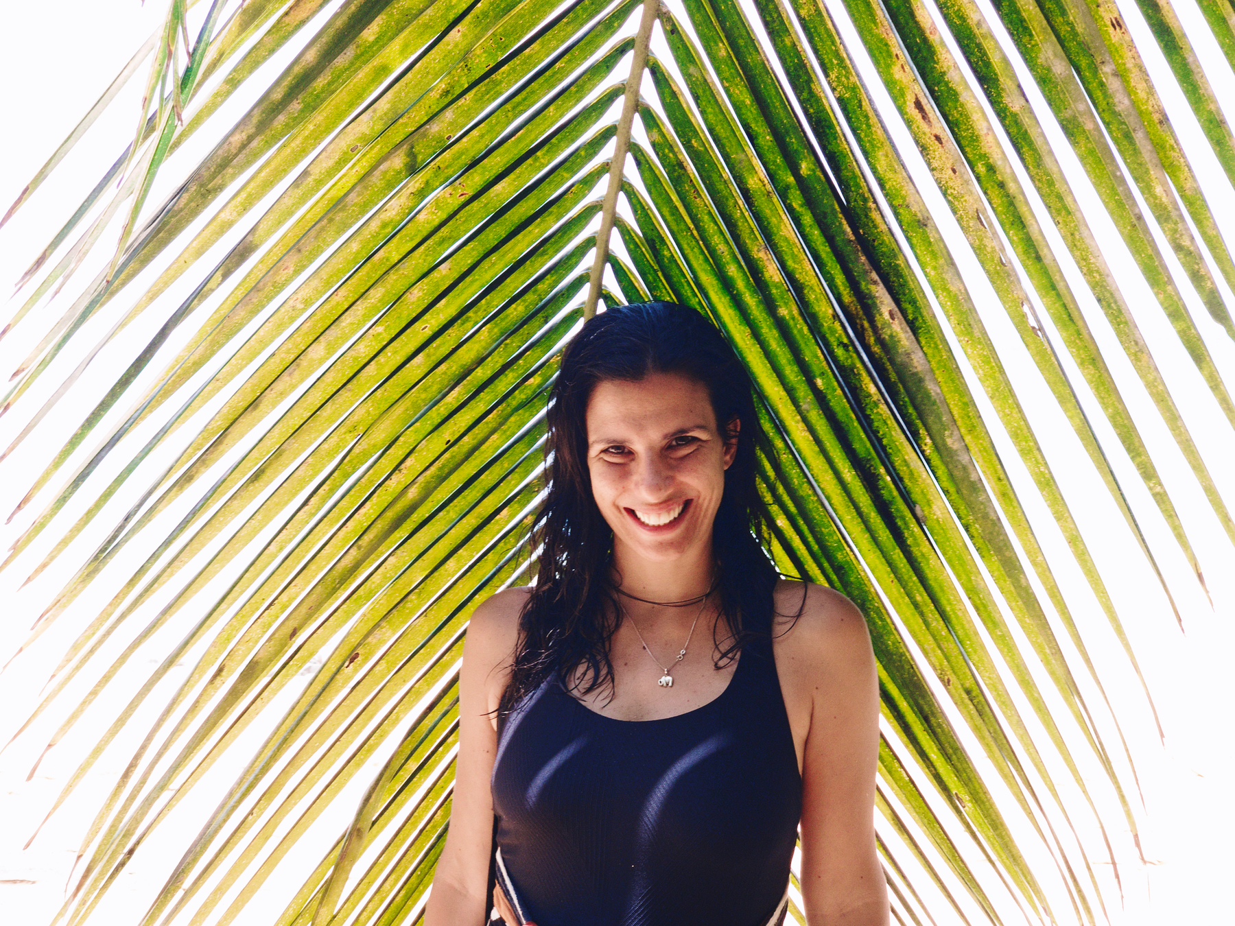 A woman looks at us, smiling, with a palm tree branch behind her. 