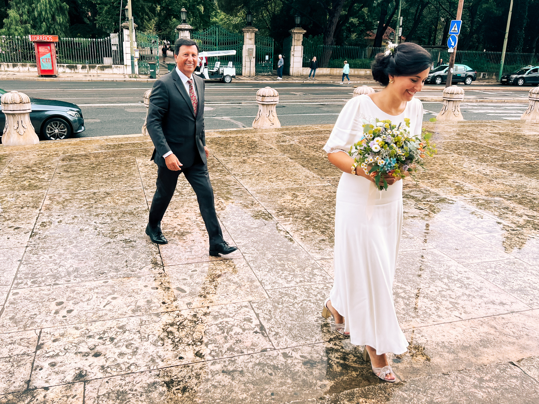 A bride walks towards the church. Her happy father follows close behind. 