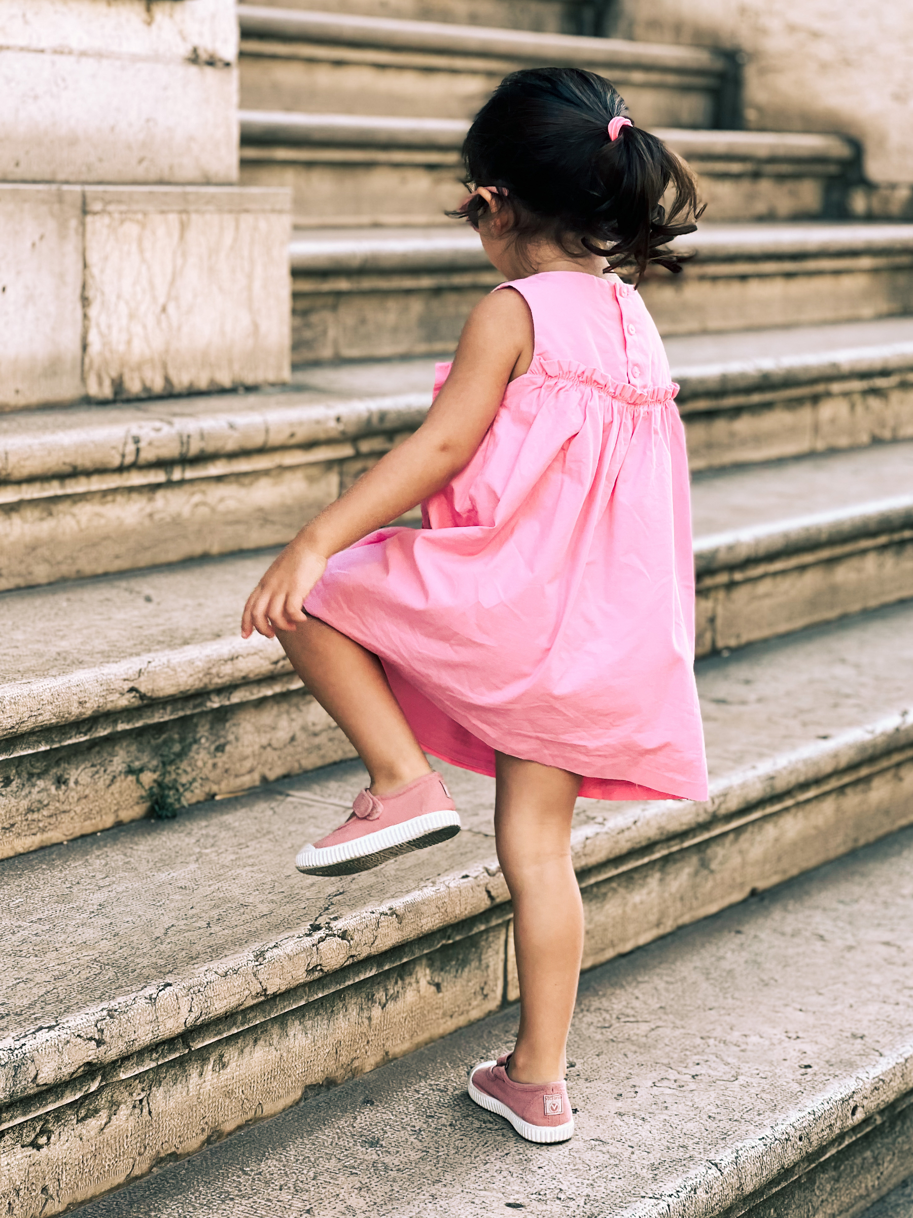 A girl wearing a pink dress walks up stone stairs. 