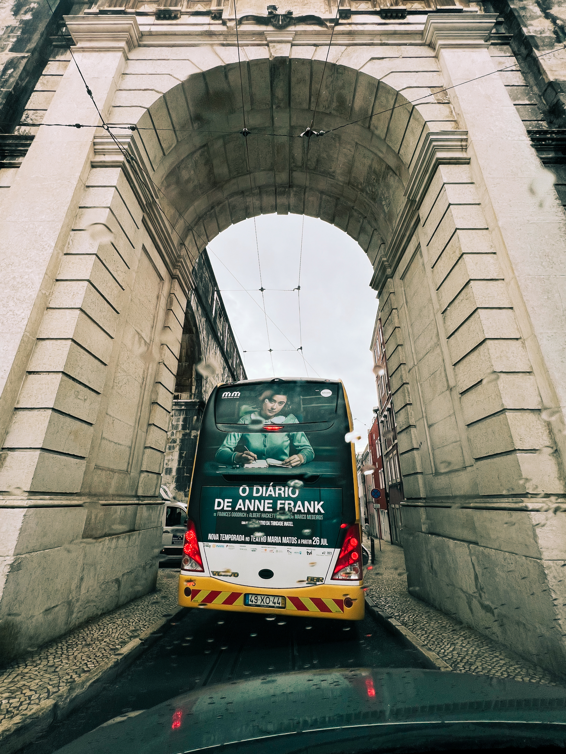 A bus is stopped under an aqueduct arch. There’s an ad for a theater play, “Diary of Anne Frank”. It’s raining. 