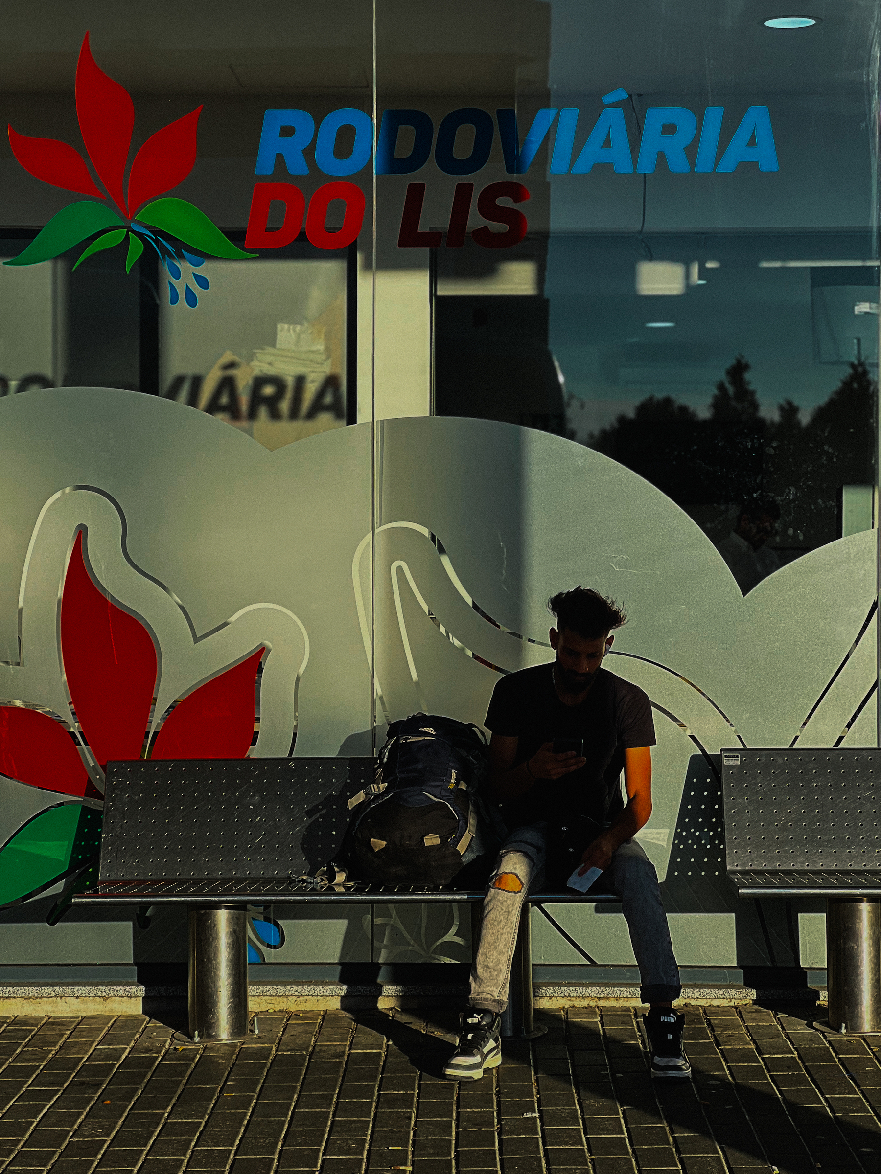 A man sits in the shade of a column, on a metal bench. Bus depot. 