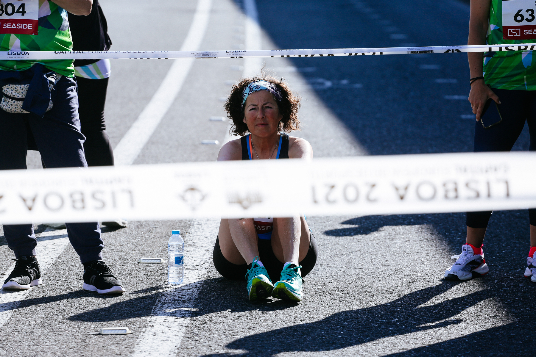 A woman sits on the pavement. She’s a runner, relaxing before a race. On both sides we can see other runners. 