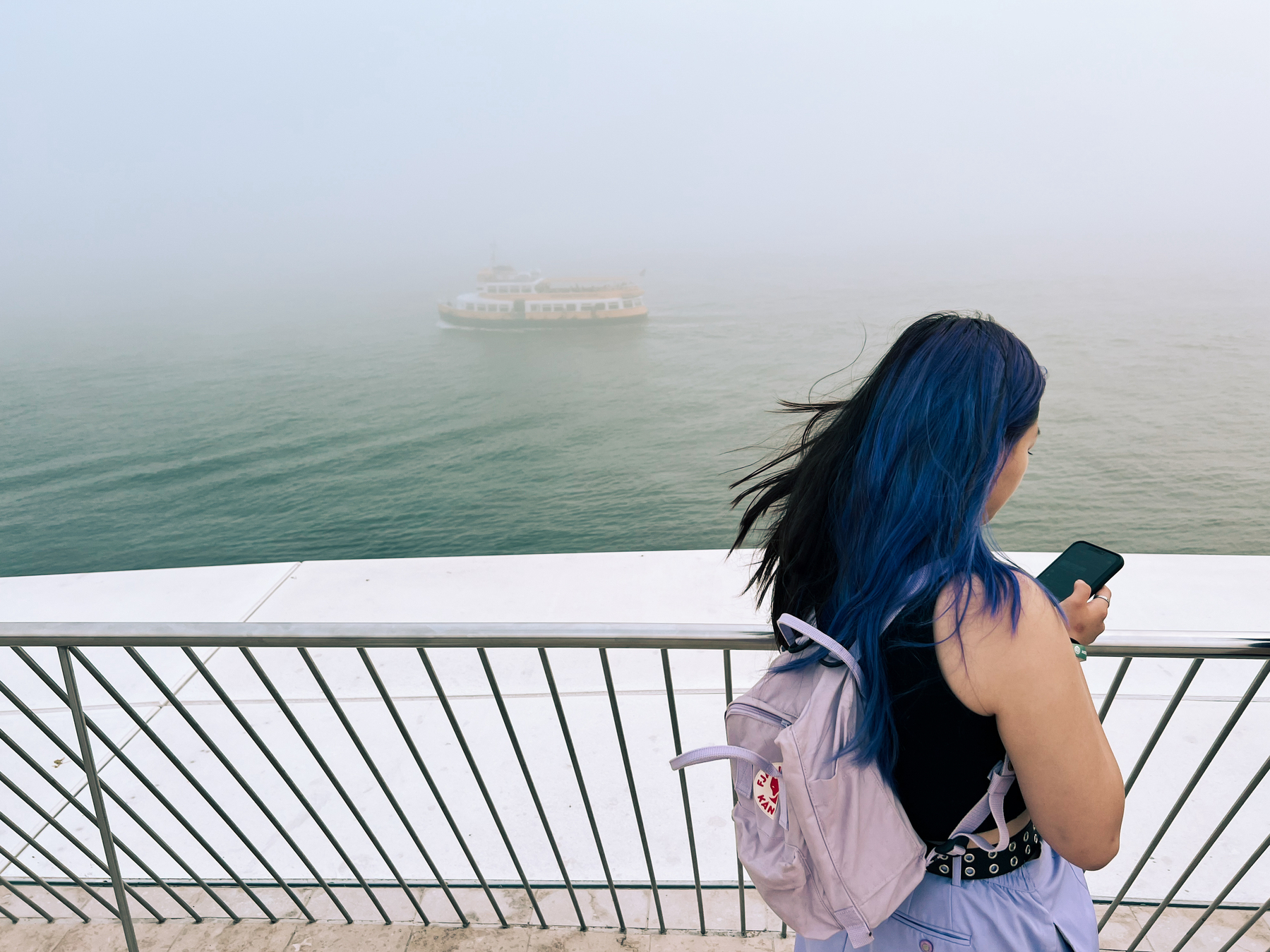 A girl checks her phone, while a ferry sails by. Foggy. 