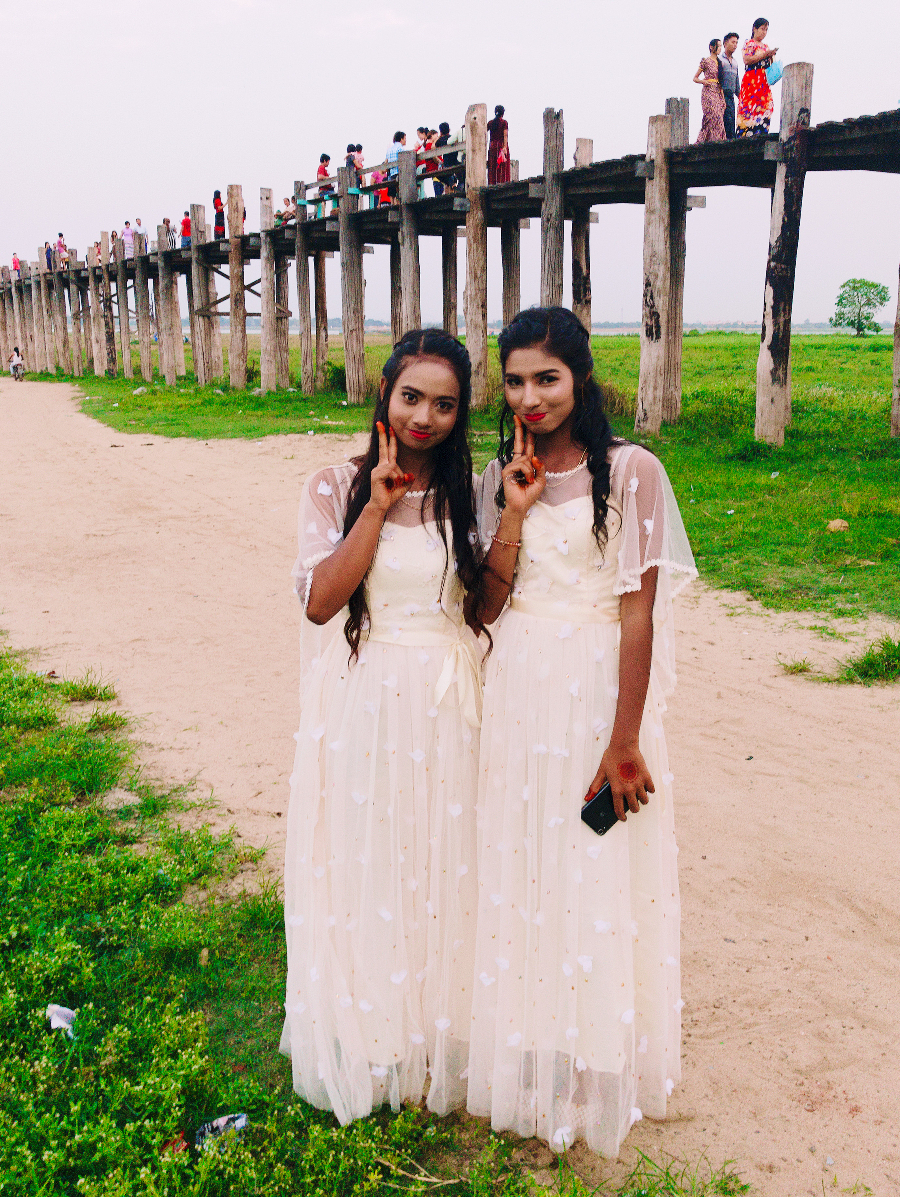 Two girls dressed in white dresses look at the cameras, flashing “V” signs with their hands. A bridge with people going by on the background. 