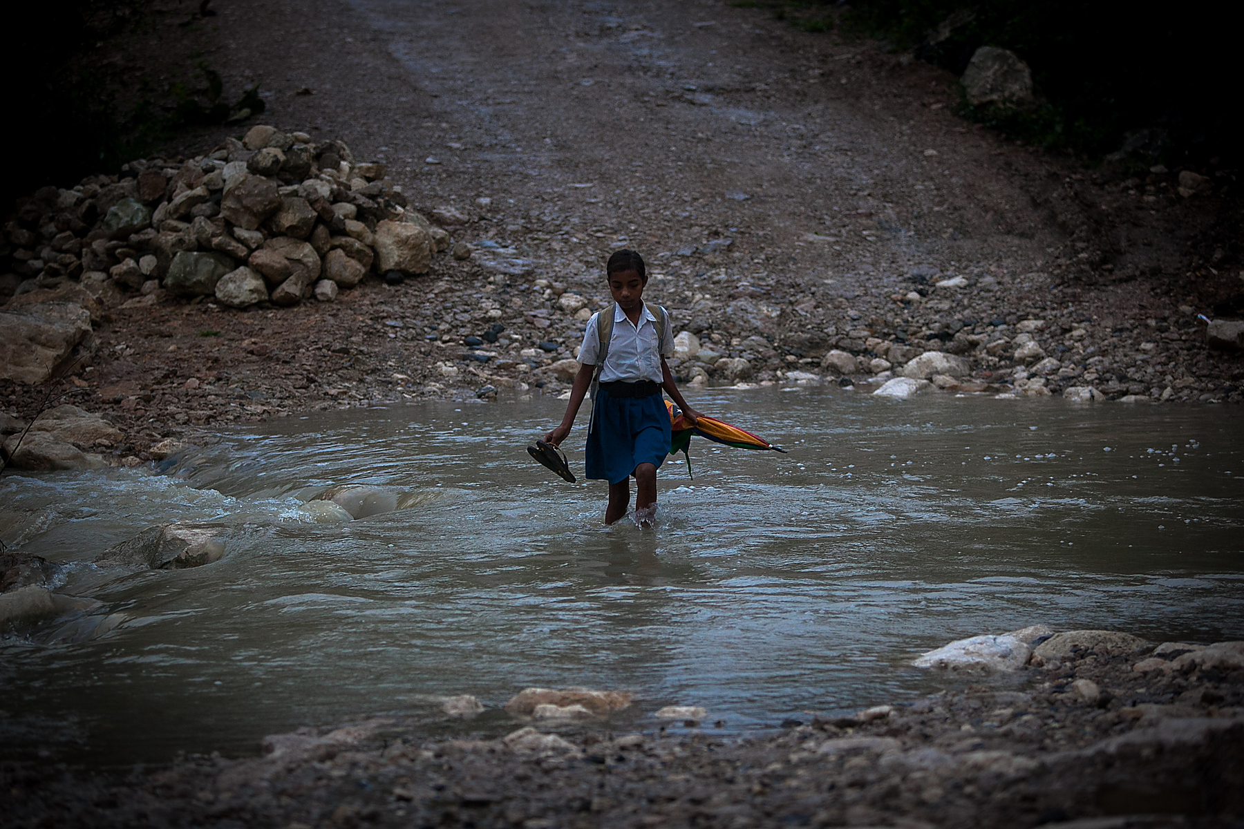 A girl crossing a river on foot, holding an umbrella, and her shoes.