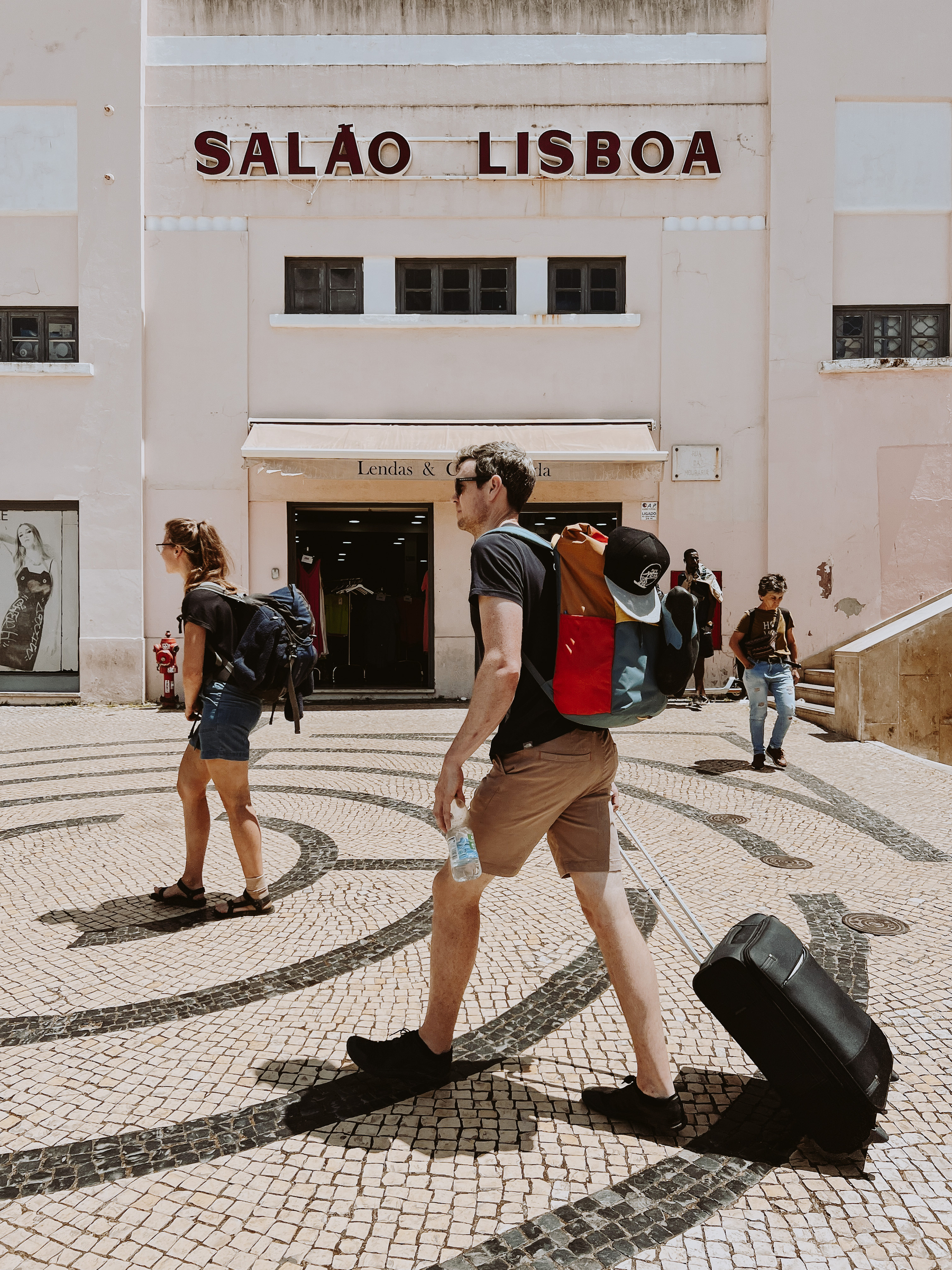 Tourists walk by, in front of a building that has the words “Salão Lisboa” on the facade. Both have backpacks, one ia also pulling a trolley. 