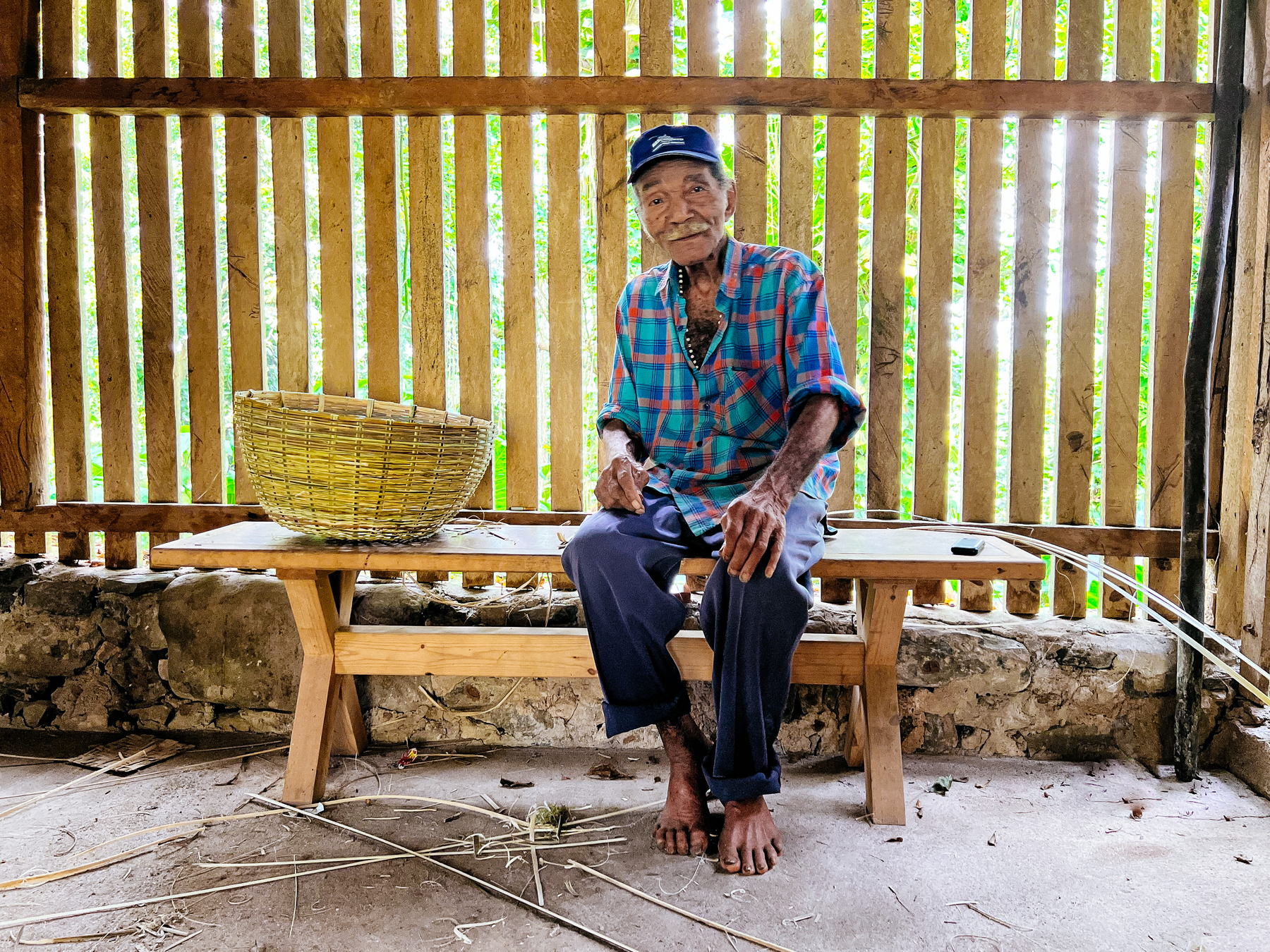 A man sits in a bench, with a basket by his side. He’s a basket weaver. 